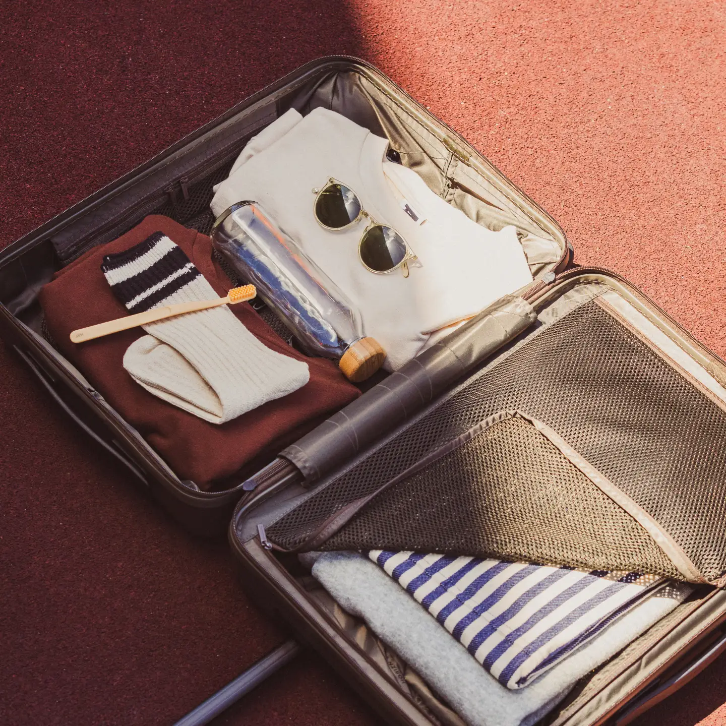 An open suitcase lies on the floor, containing a toothbrush, socks, sunglasses, an empty water bottle and a few items of clothing.