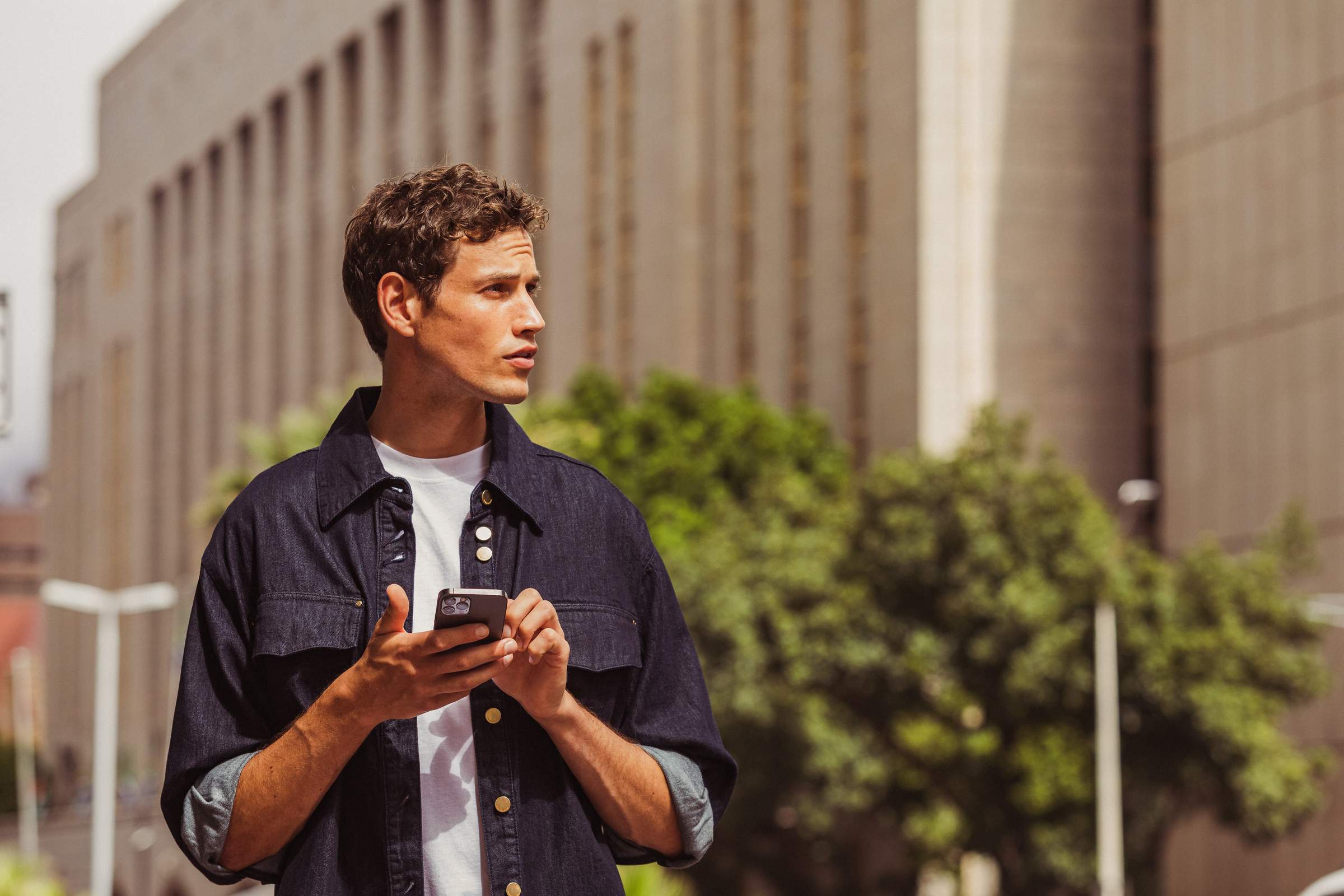 Un joven con una camisa oscura está de pie con su smartphone en un lado de la calle en una ciudad. Al fondo se ven un árbol y un edificio alto. El sol brilla.