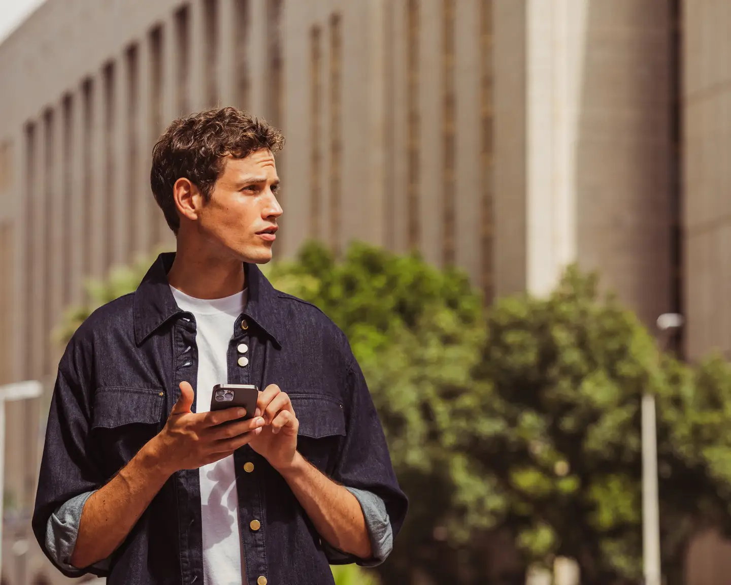 Un joven con una camisa oscura está de pie con su smartphone en un lado de la calle en una ciudad. Al fondo se ven un árbol y un edificio alto. El sol brilla.