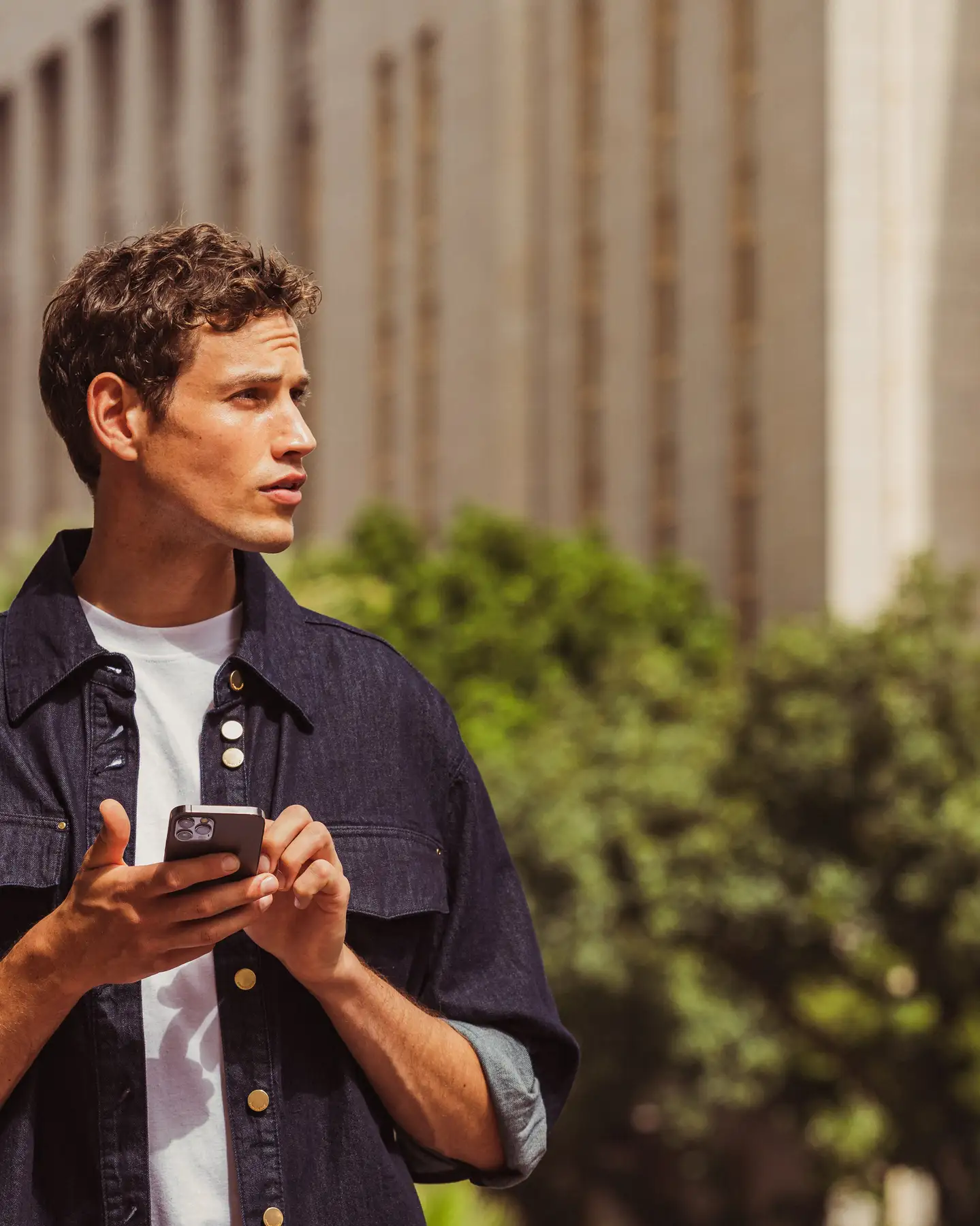Un joven con una camisa oscura está de pie con su smartphone en un lado de la calle en una ciudad. Al fondo se ven un árbol y un edificio alto. El sol brilla.