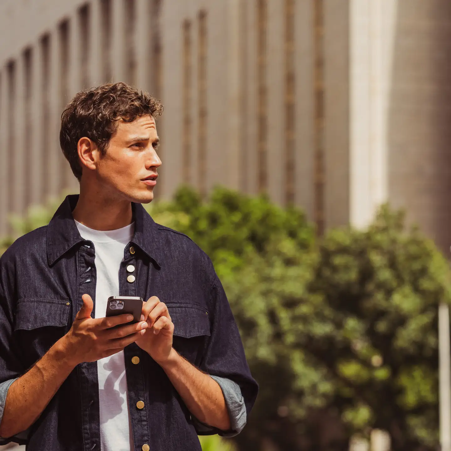 Un joven con una camisa oscura está de pie con su smartphone en un lado de la calle en una ciudad. Al fondo se ven un árbol y un edificio alto. El sol brilla.