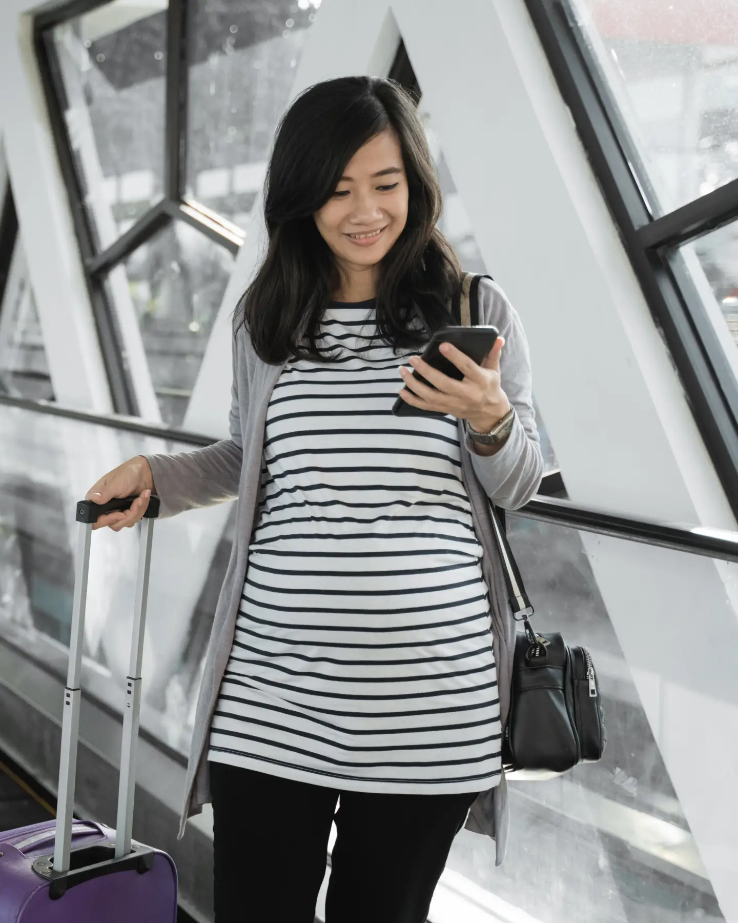 Een zwangere vrouw staat op de luchthaven met haar handbagage en kijkt naar haar smartphone.