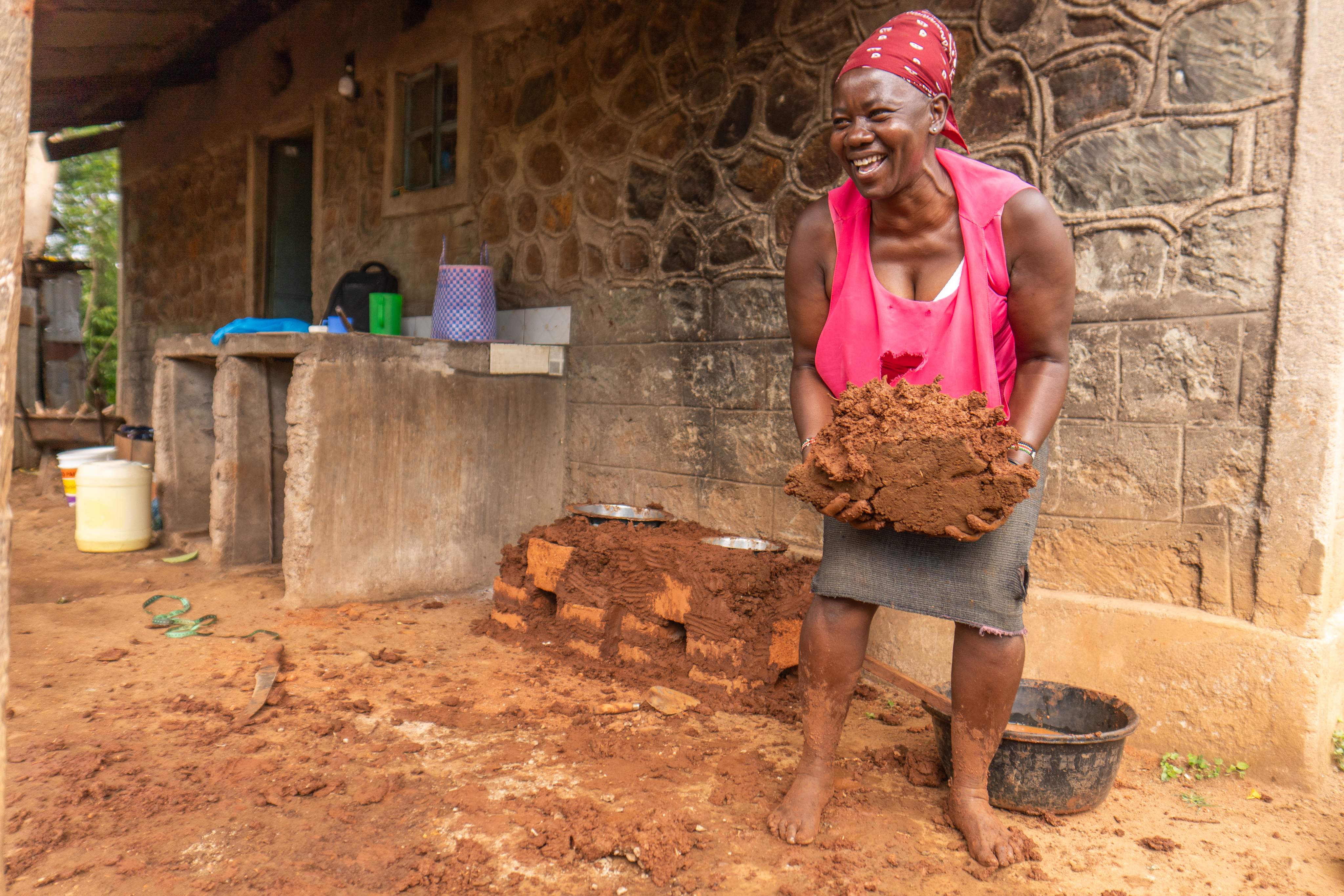 Een lachende vrouw met een hoofddoek houdt natte klei in haar handen terwijl ze voor een stenen gebouw staat. Op de achtergrond zijn eenvoudige huishoudelijke items te zien.
