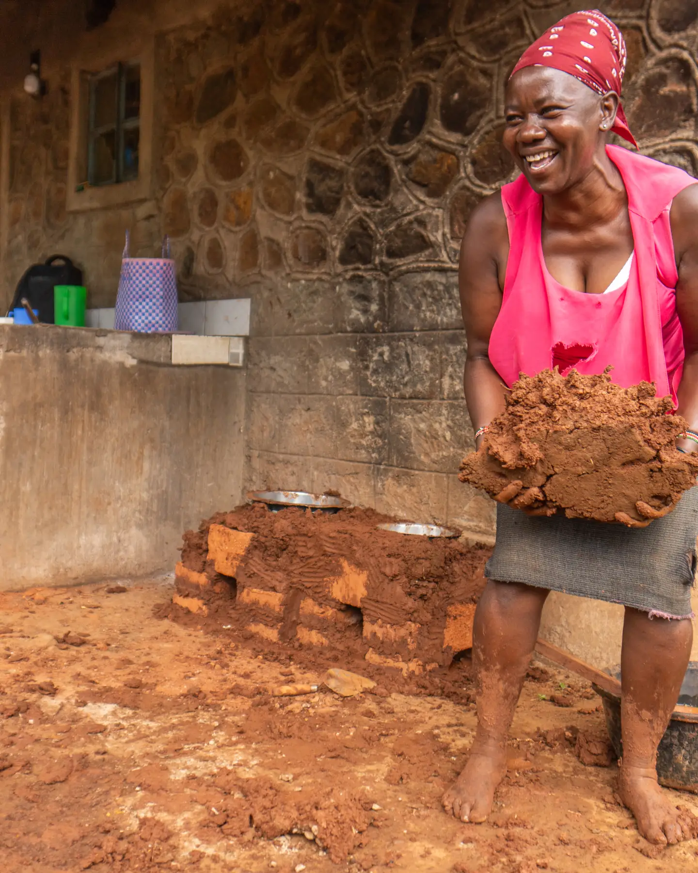 Une femme souriante, coiffée d'un foulard, tient de l'argile humide dans ses mains devant un bâtiment en pierre. Des objets ménagers simples sont visibles à l'arrière-plan.