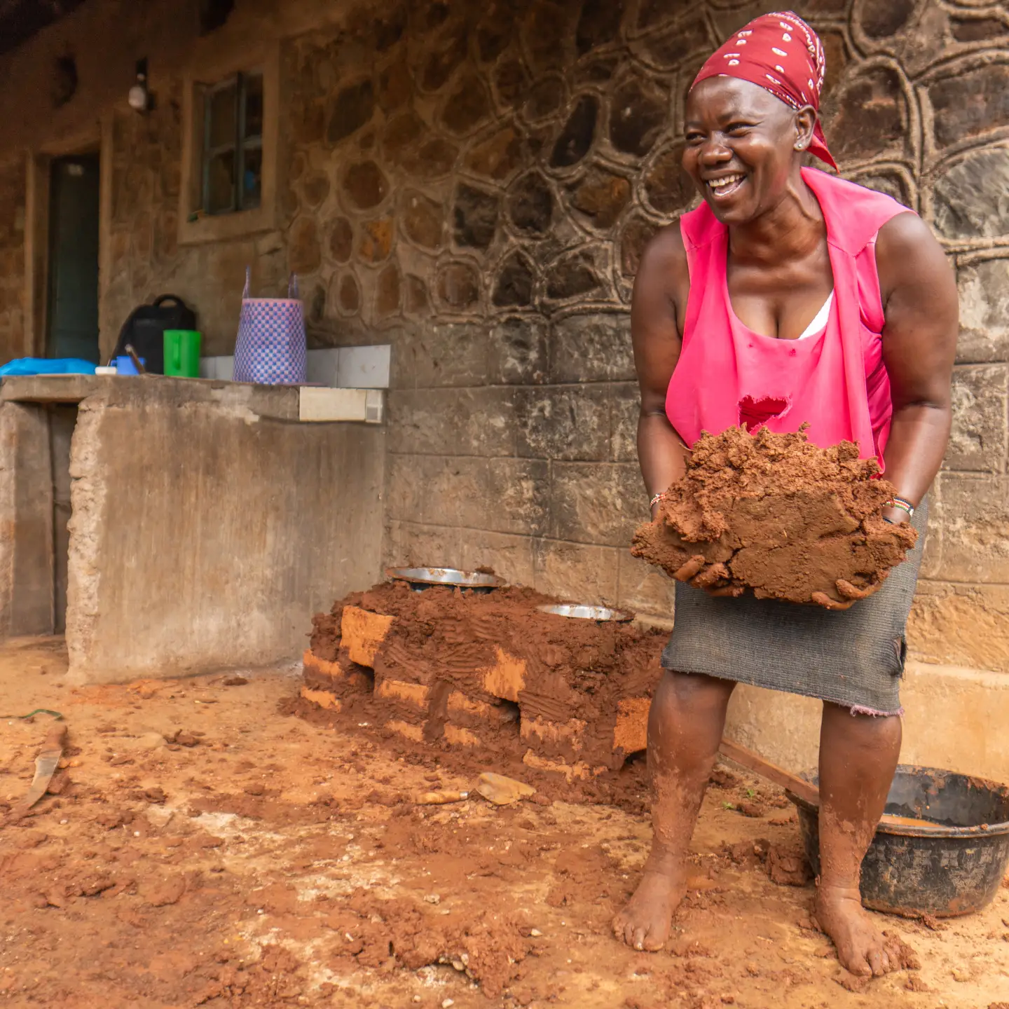 Une femme souriante, coiffée d'un foulard, tient de l'argile humide dans ses mains devant un bâtiment en pierre. Des objets ménagers simples sont visibles à l'arrière-plan.