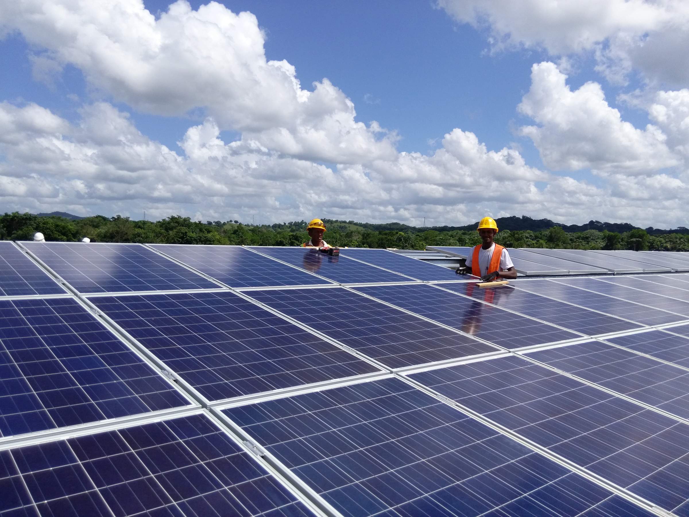 Dos trabajadores con chalecos naranjas y cascos amarillos instalan paneles solares en un tejado bajo un cielo soleado con nubes blancas, árboles verdes y colinas al fondo.