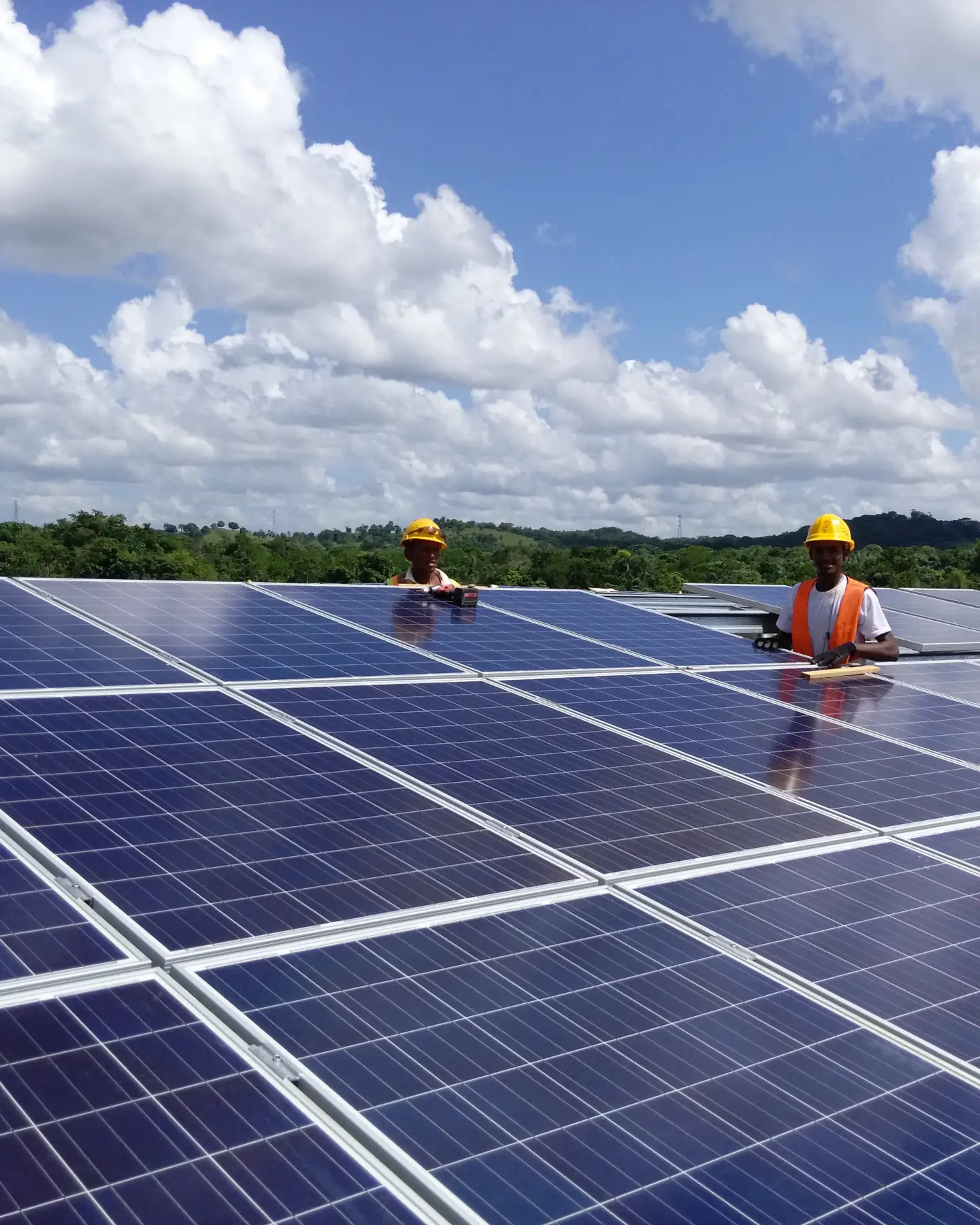 Twee mannen met oranje hesjes en gele helmen zijn bezig met het plaatsen van zonnepanelen op een dak. De zon schijnt, er zijn wat witte wolken, en op de achtergrond zijn groene bomen en heuvels te zien.