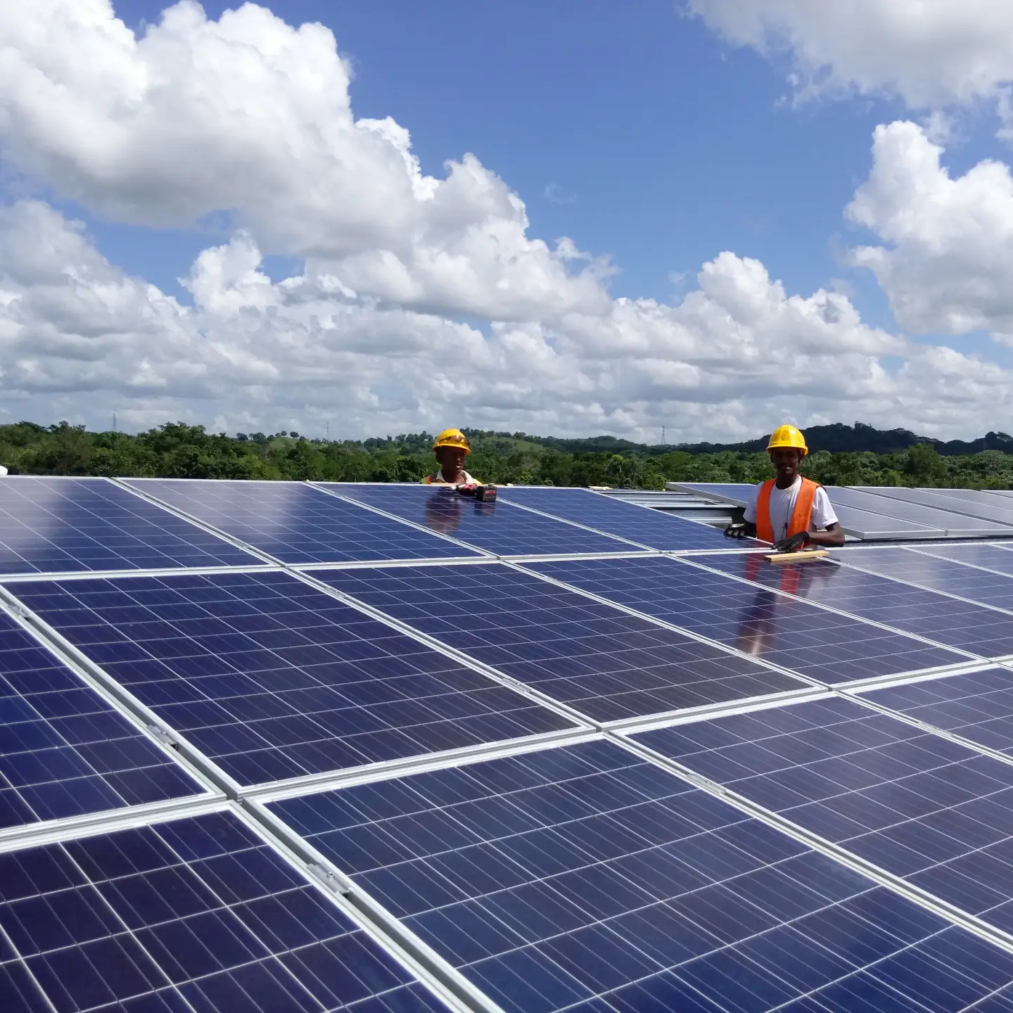 Sous un ciel ensoleillé et nuageux, deux travailleurs en gilets orange et casques jaunes posent des panneaux solaires sur un toit, entourés d’arbres verdoyants et de collines en toile de fond.