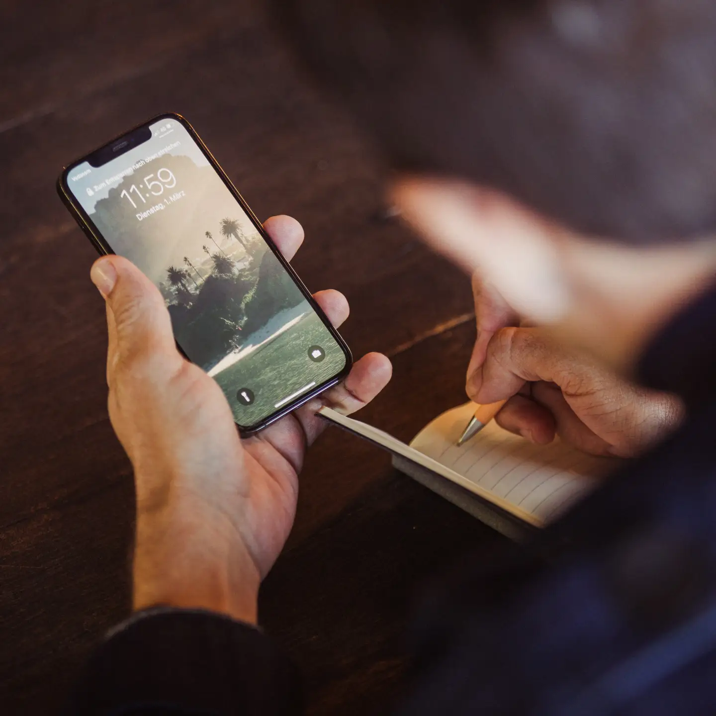 Un hombre sostiene un smartphone y escribe algo en su cuaderno al mismo tiempo.
