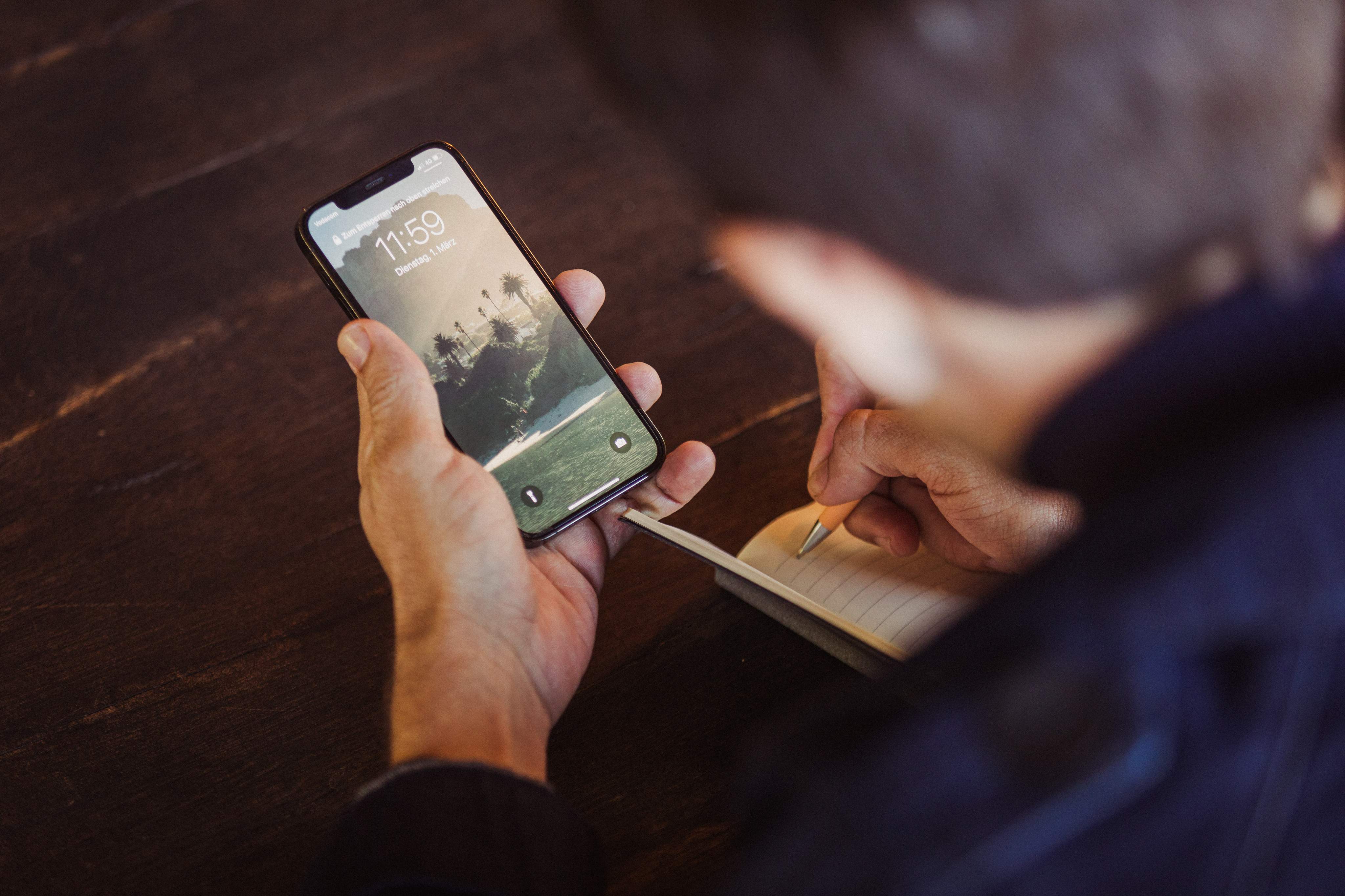 Un hombre sostiene un smartphone y escribe algo en su cuaderno al mismo tiempo.