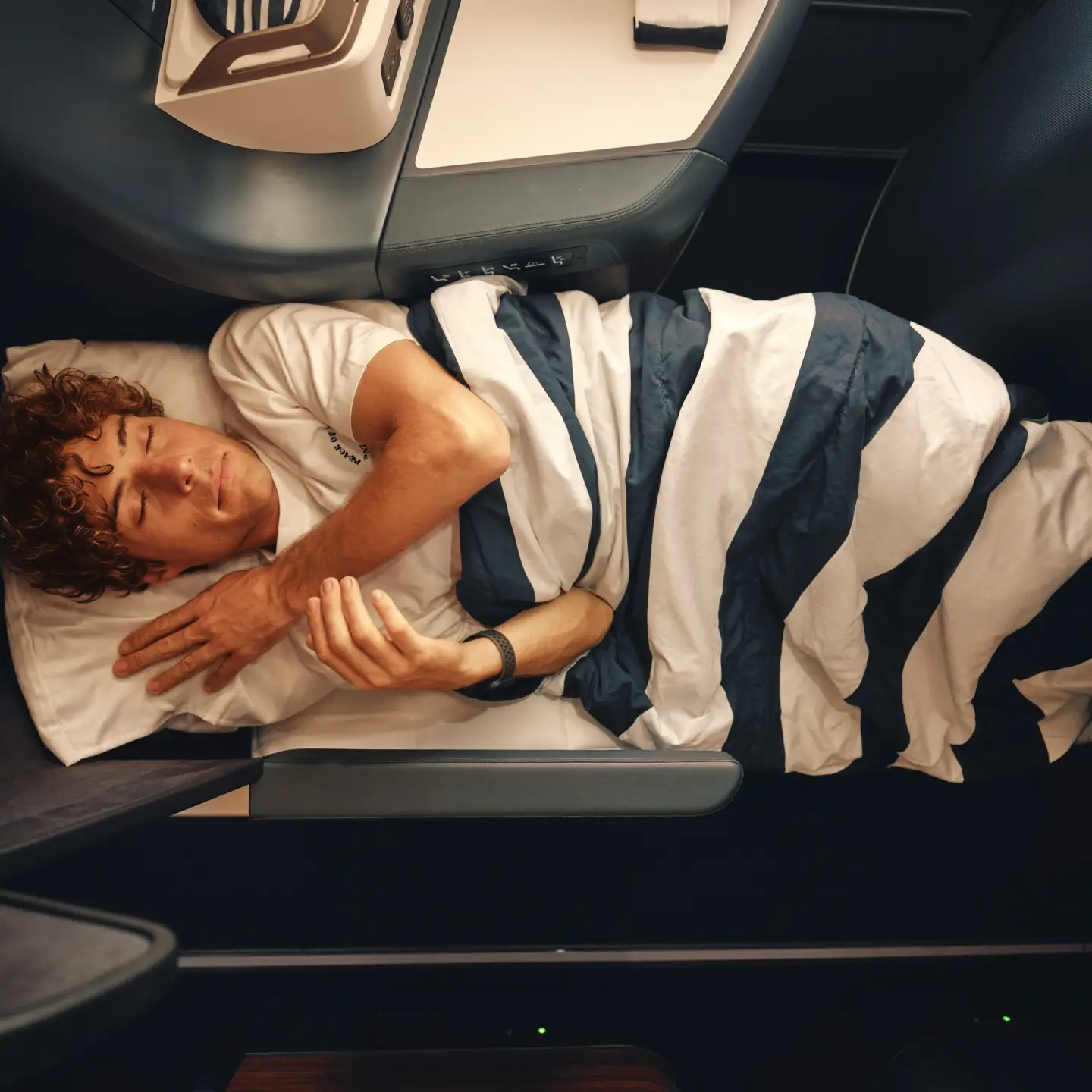 Un hombre duerme bajo una manta a rayas en un asiento de Business Class