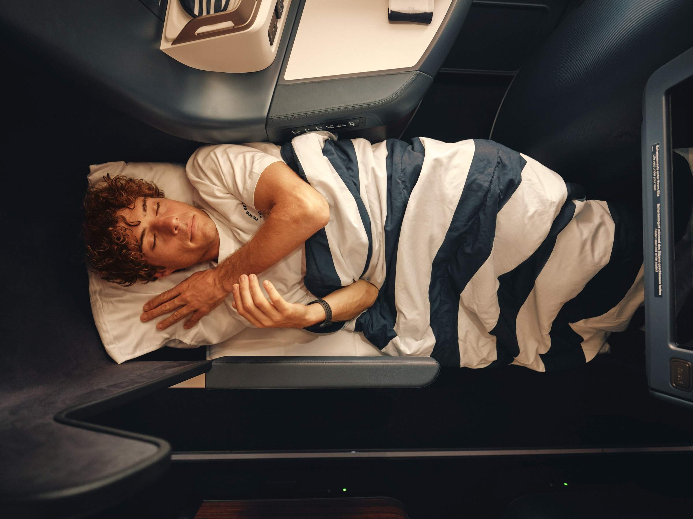 Un hombre duerme bajo una manta a rayas en un asiento de Business Class 