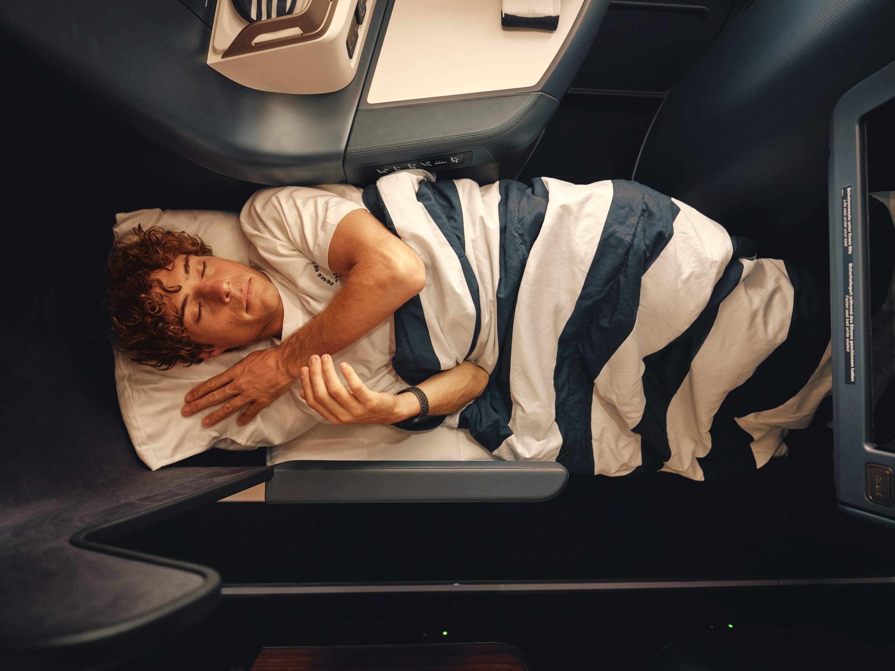 Een man slaapt onder een gestreepte deken op een stoel in de Business Class. 