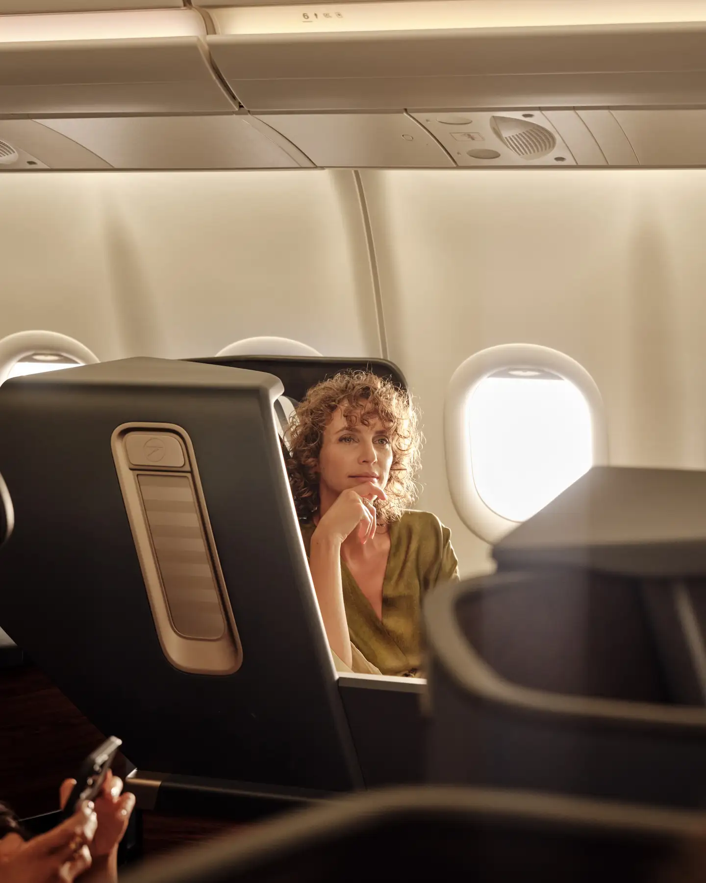 Una mujer se sienta pensativa en el asiento de Business Class del avión mientras mira por la ventana, rodeada de una suave luz solar.