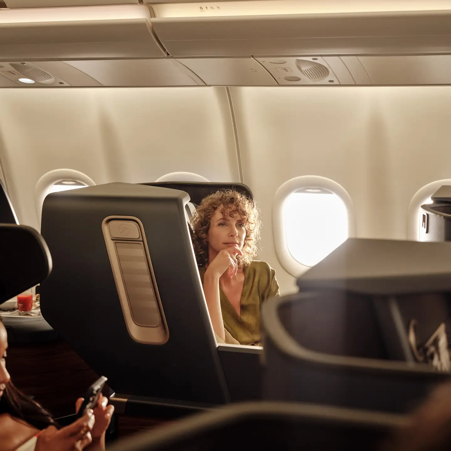 Una mujer se sienta pensativa en el asiento de Business Class del avión mientras mira por la ventana, rodeada de una suave luz solar.