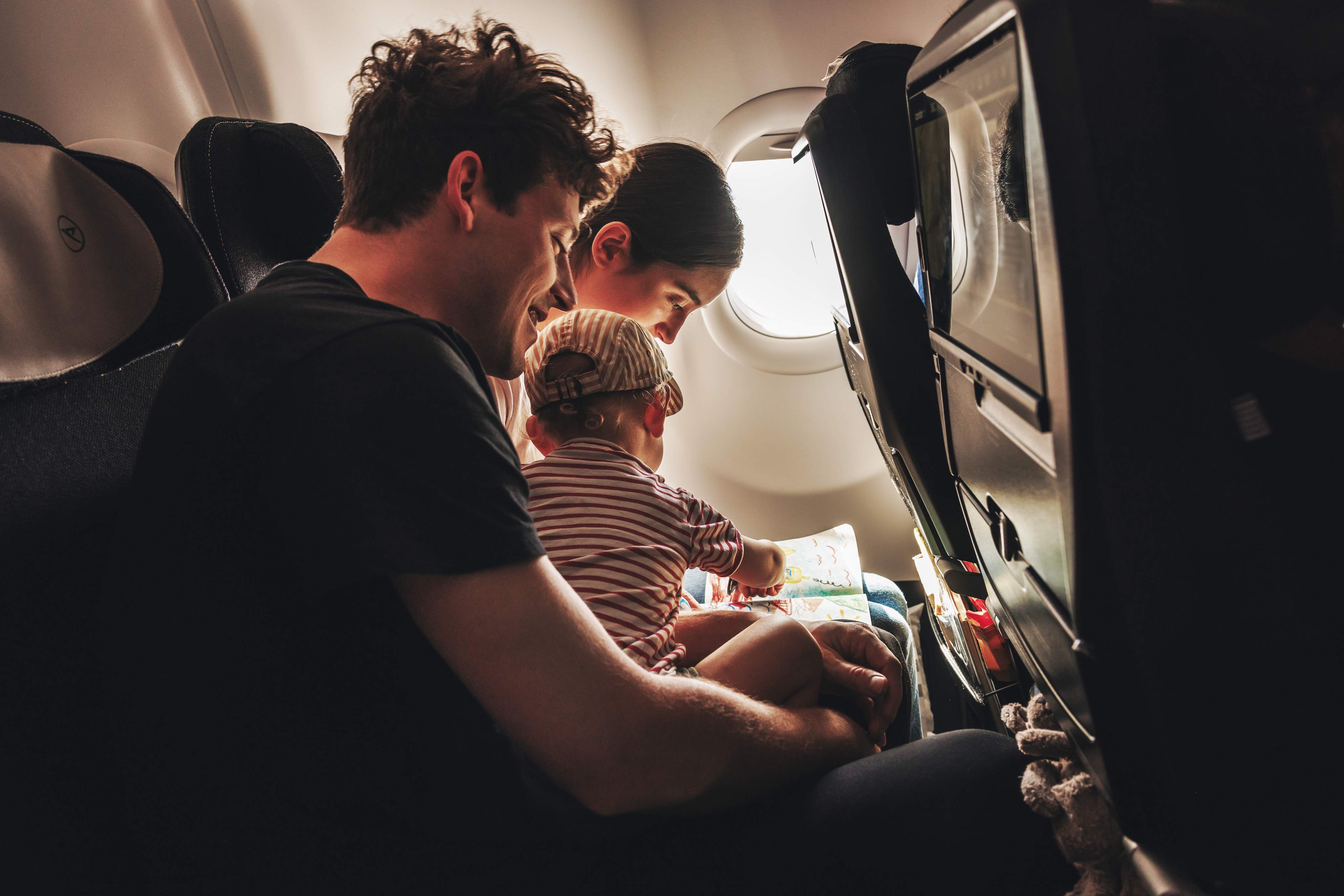 Une jeune famille, composée d'un homme, d'une femme et d'un bébé portant un t-shirt et une casquette rayés rouges, assise dans la cabine d'un Airbus A330neo. Le bébé est assis sur les genoux de son père et dessine sur un livre à colorier.