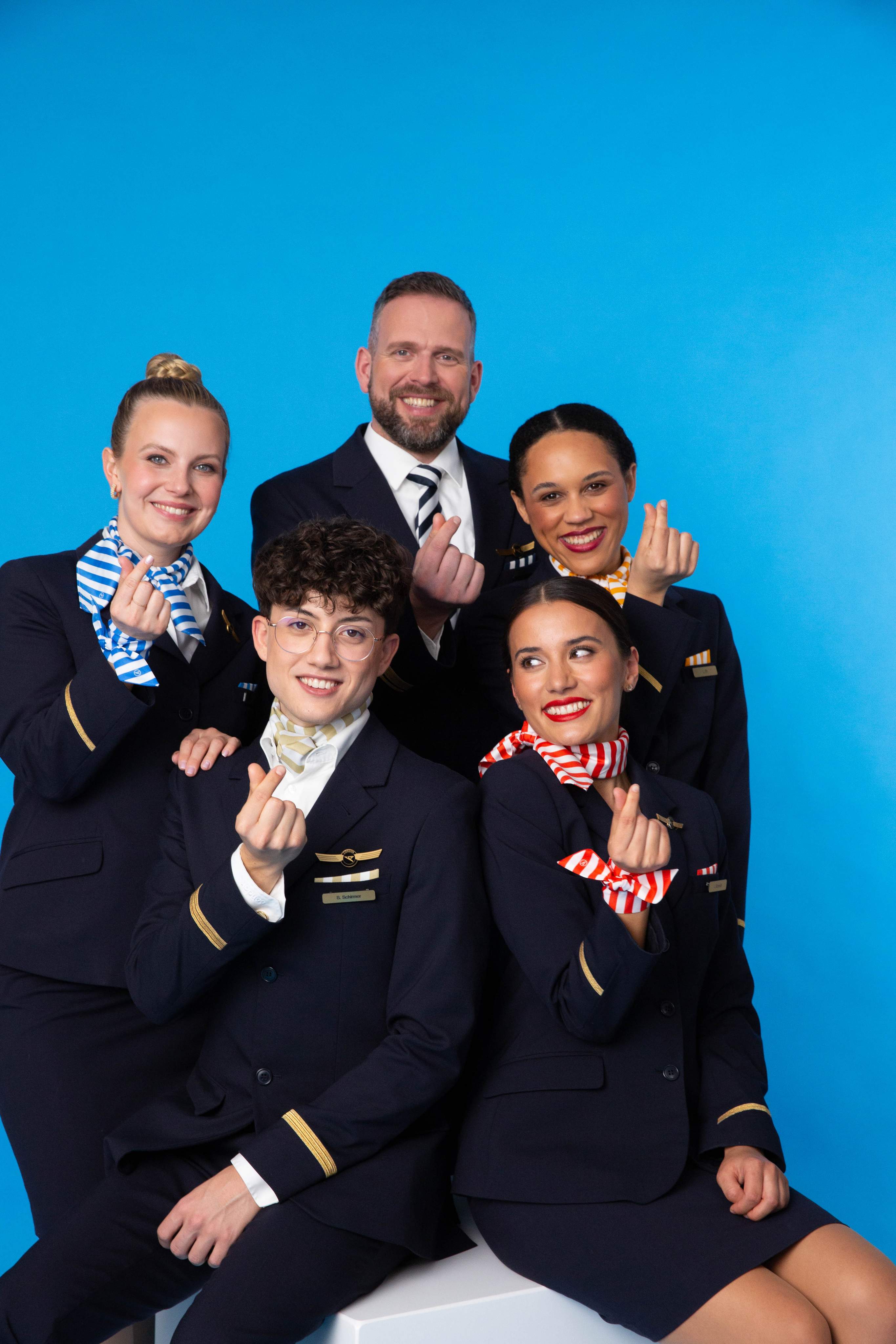 Condor-bemanning met stewardessen, purser en piloot voor een zeeblauwe achtergrond, in uniform en met gestreepte accessoires.