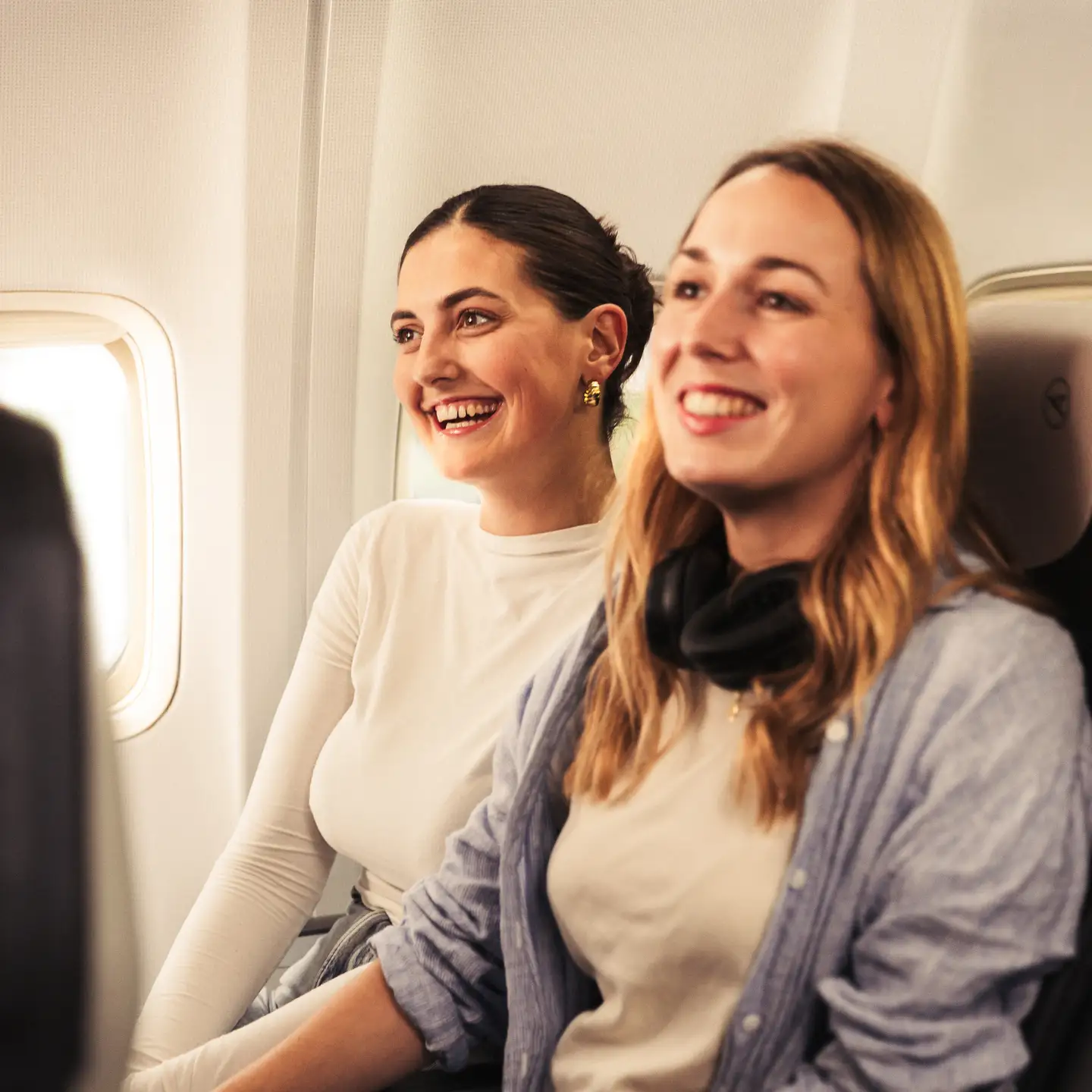 Dos mujeres jóvenes sentadas una junto a la otra en asientos azul oscuro en un avión, ambas sonriendo