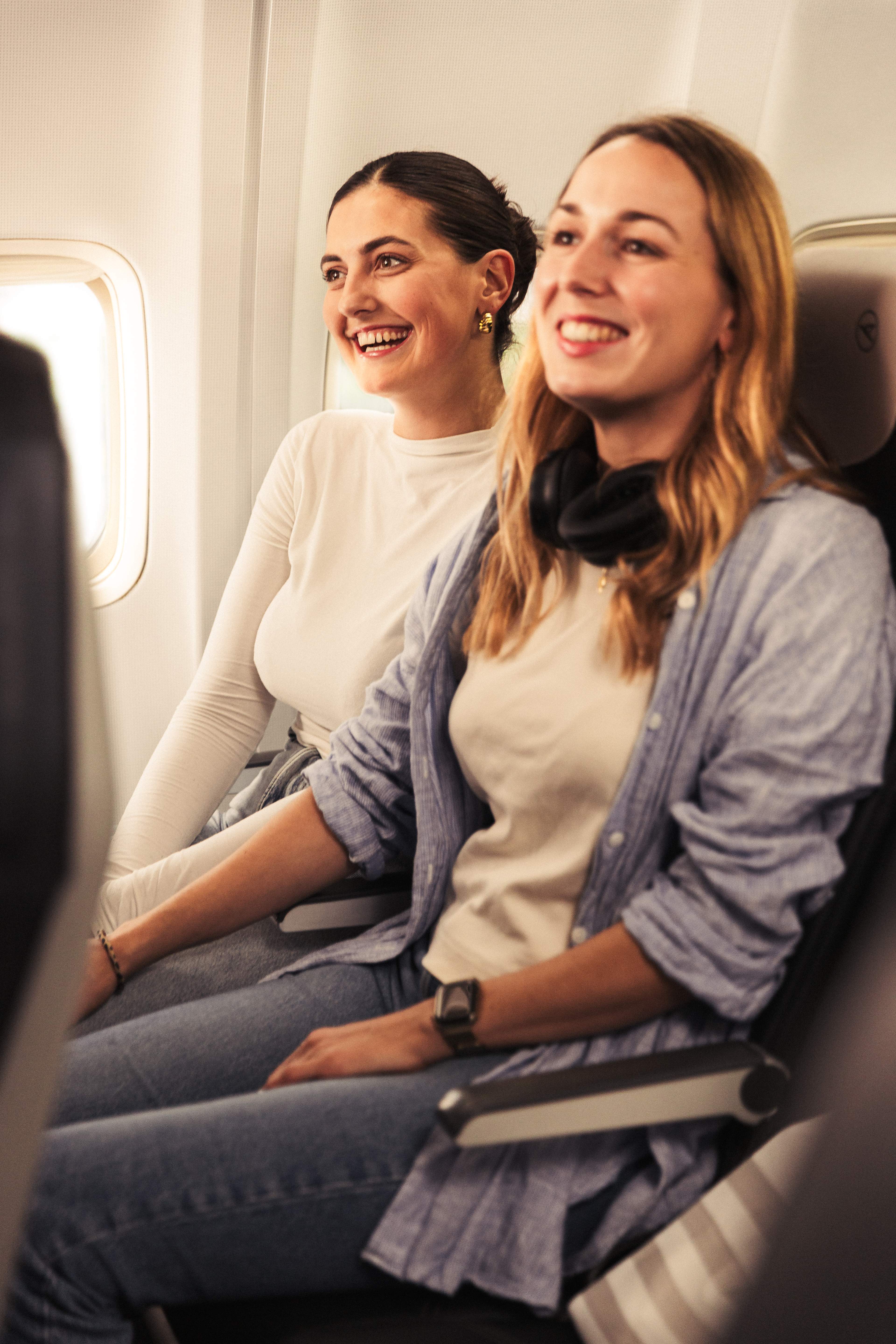 Deux jeunes femmes assises côte à côte sur des sièges bleu foncé dans un avion, toutes deux souriantes