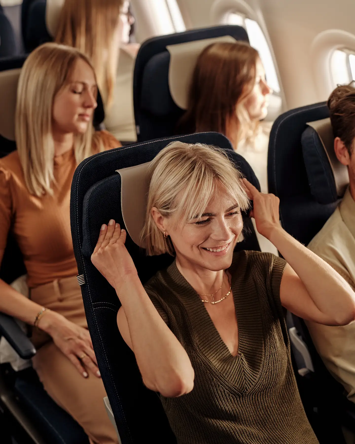 Una mujer sonriente se relaja en un avión y ajusta su reposacabezas, mientras otros pasajeros permanecen sentados y dormidos al fondo.