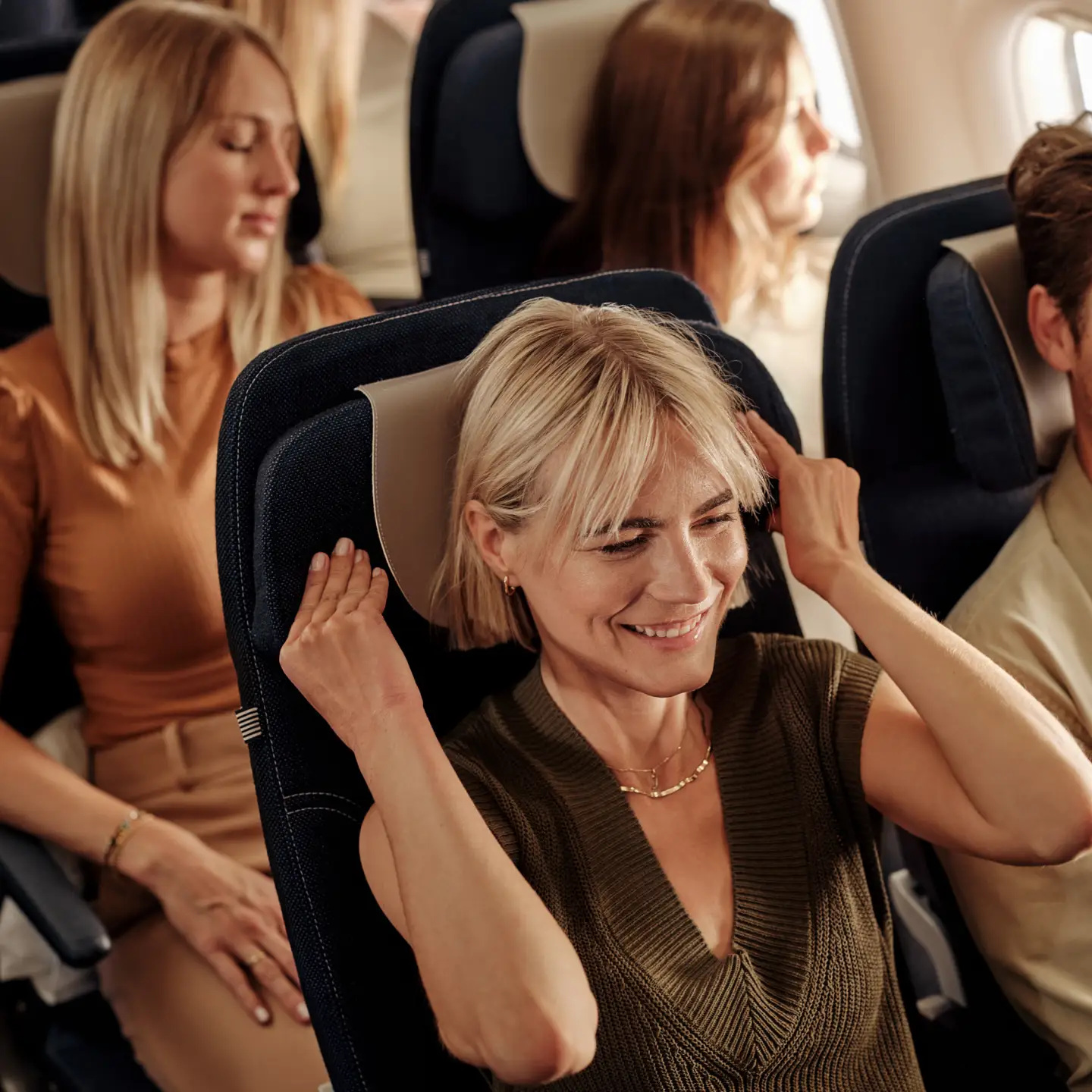 A smiling woman relaxing on a plane, adjusting her headrest as other passengers sit and doze in the background.