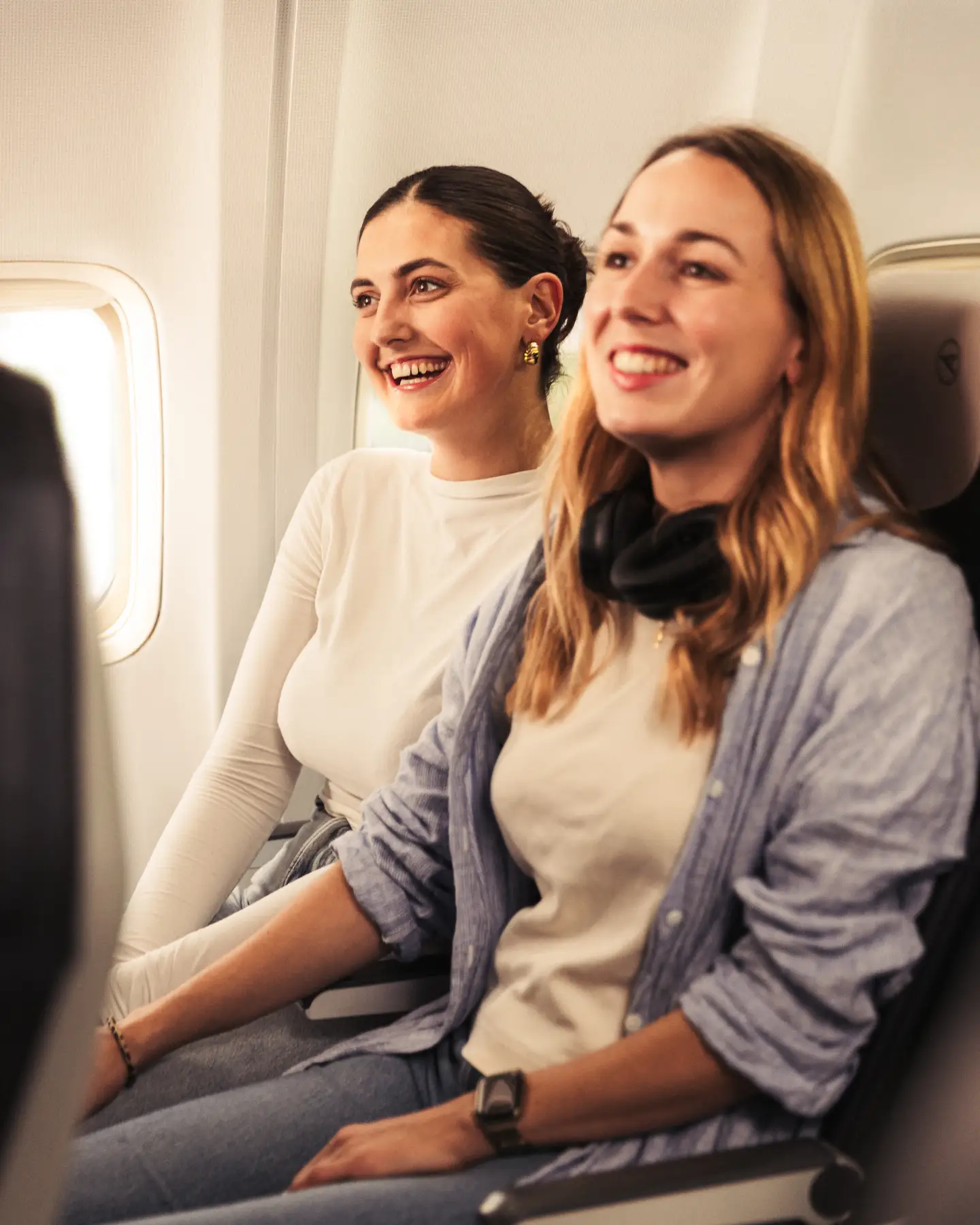 Two young women sitting next to each other on dark blue seats on a plane, both smiling
