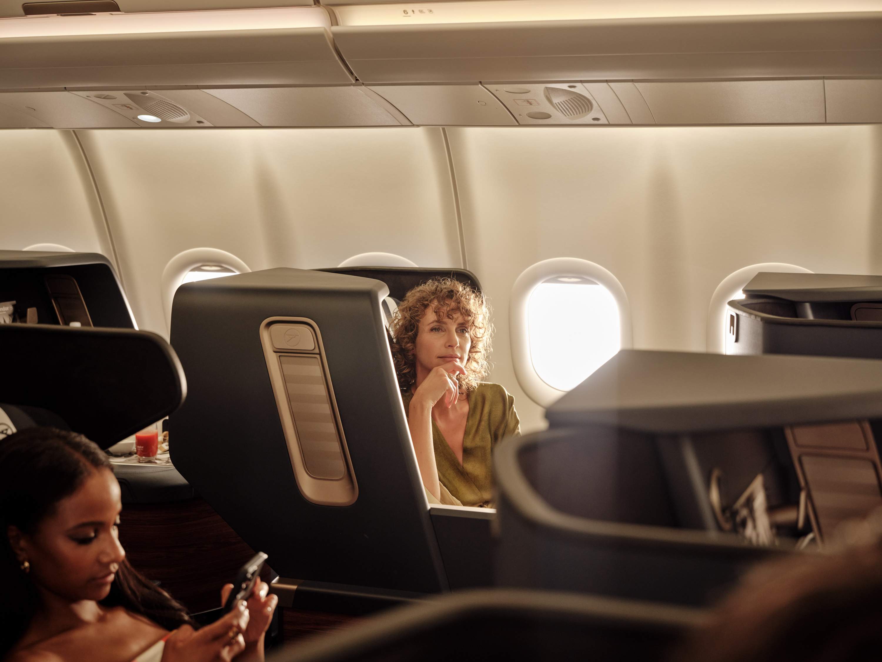 Žena zamyšleně sedící na sedadle Business Class v letadle a hledící z okna, obklopená jemným slunečním světlem.