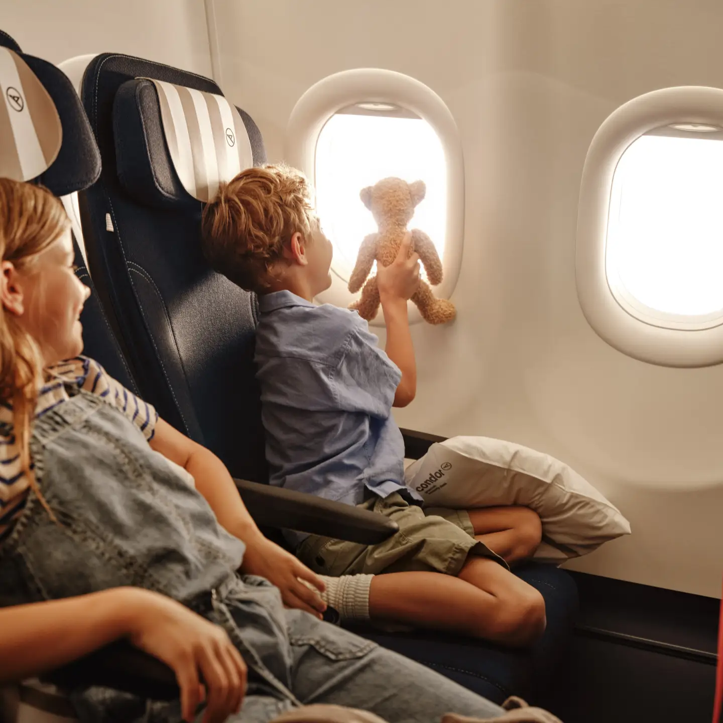 Twee kinderen zitten aan boord van een Condor-vliegtuig, één van hen kijkt uit het raam.