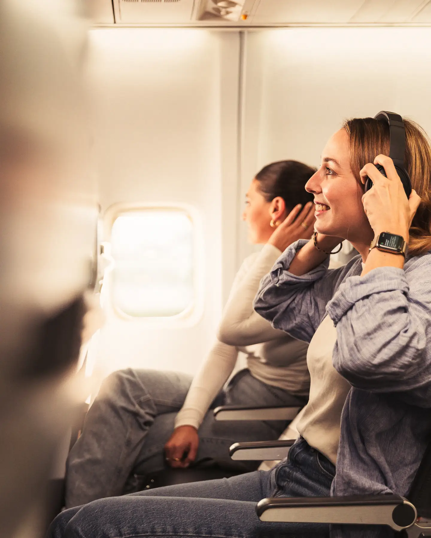 Two women are sitting next to each other in a plane. The woman in the foreground is wearing headphones, smiling and looking out of the window, while the other woman, also wearing headphones, is leaning back and relaxing.