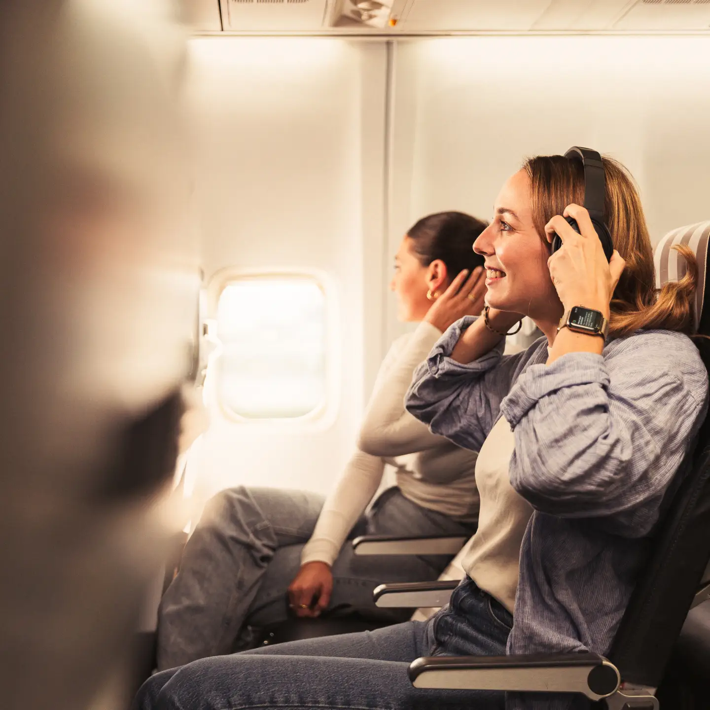 Two women are sitting next to each other in a plane. The woman in the foreground is wearing headphones, smiling and looking out of the window, while the other woman, also wearing headphones, is leaning back and relaxing.