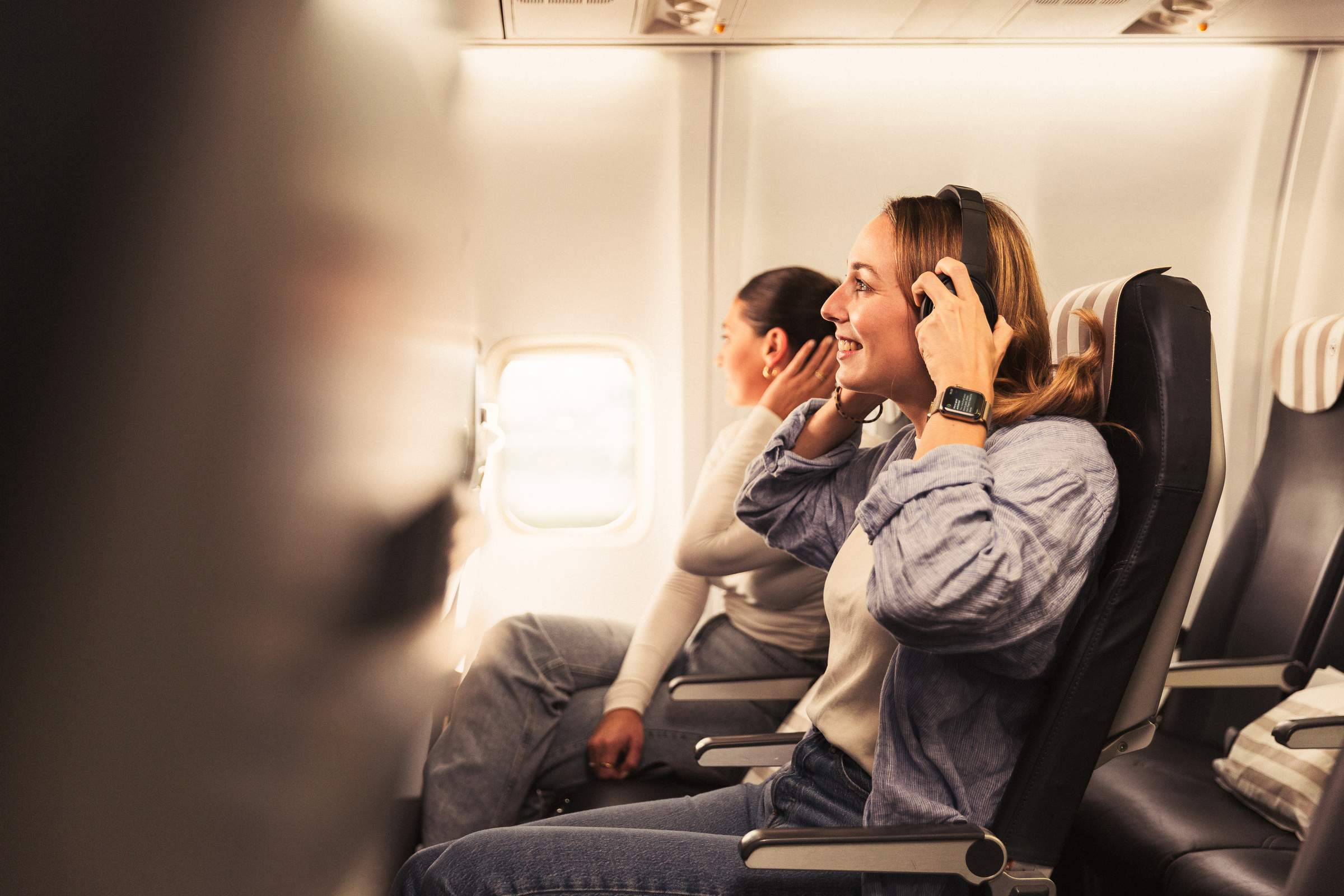 Dos mujeres están sentadas juntas en un avión. La mujer en primer plano lleva auriculares, sonríe y mira por la ventanilla, mientras que la otra mujer, también con auriculares, se reclina y se relaja.