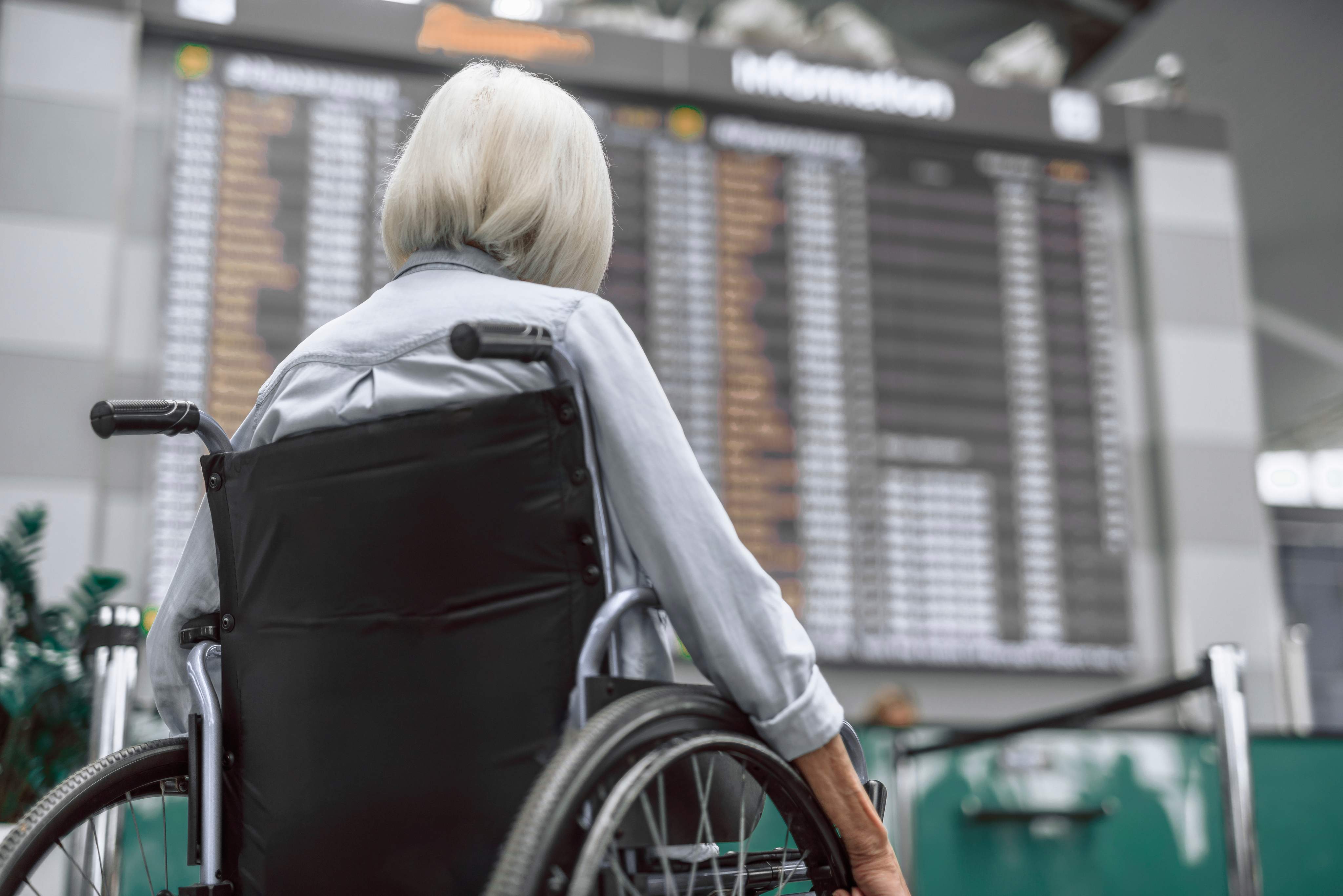 Une femme âgée aux cheveux courts et à la chemise à manches longues est assise dans un fauteuil roulant à l'aéroport devant le tableau des départs, dos à la caméra.