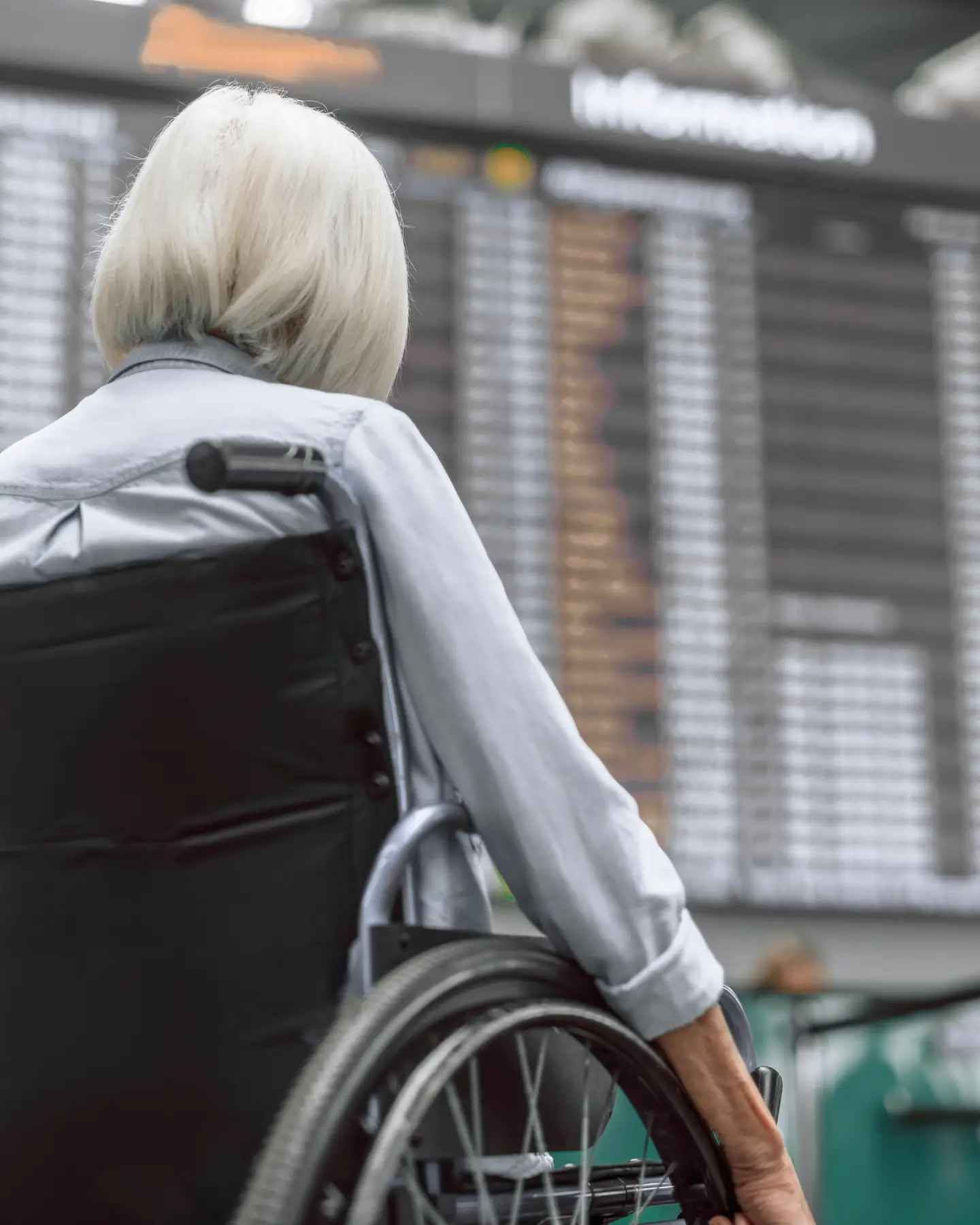 Une femme âgée aux cheveux courts et à la chemise à manches longues est assise dans un fauteuil roulant à l'aéroport devant le tableau des départs, dos à la caméra.