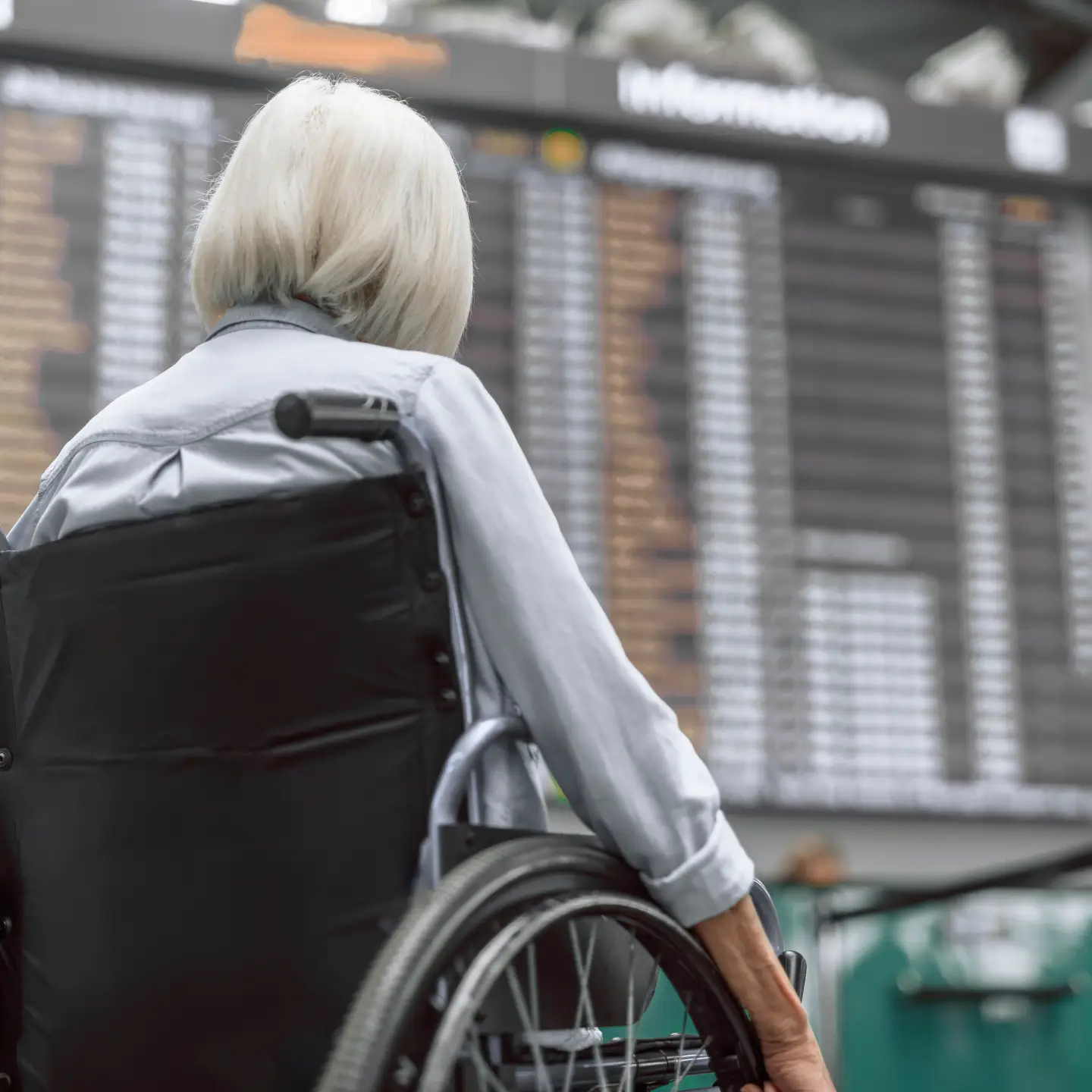 Une femme âgée aux cheveux courts et à la chemise à manches longues est assise dans un fauteuil roulant à l'aéroport devant le tableau des départs, dos à la caméra.