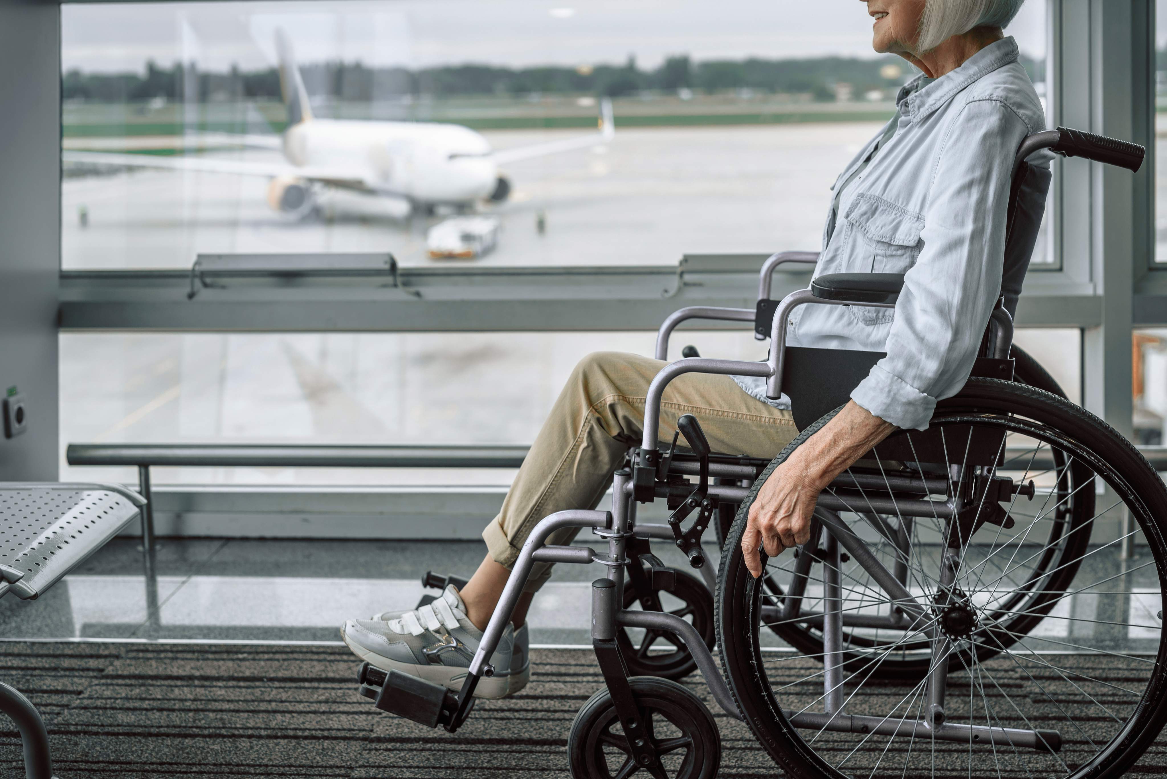 An elderly person in a wheelchair is waiting in the airport terminal, with an airplane visible on the apron in the background.