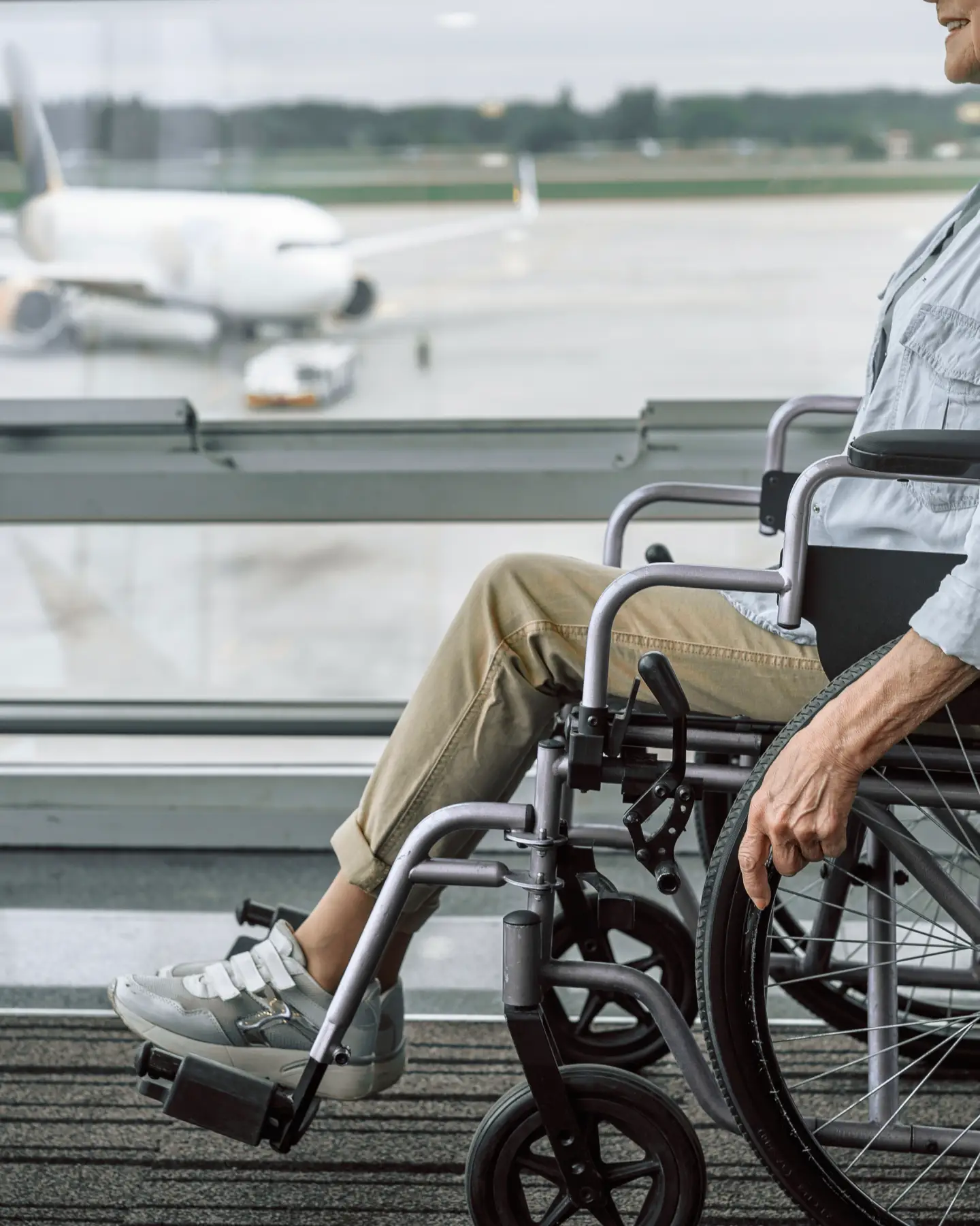 An elderly person in a wheelchair is waiting in the airport terminal, with an airplane visible on the apron in the background.