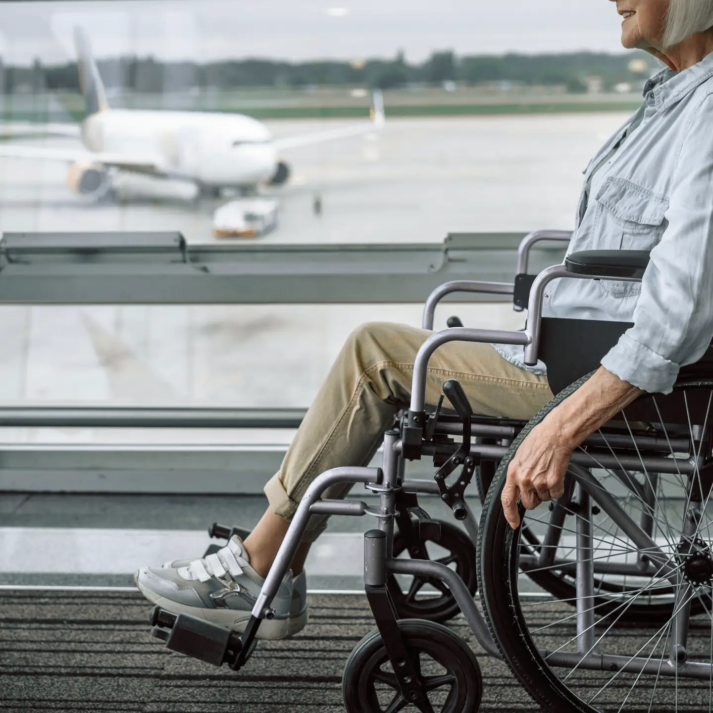 An elderly person in a wheelchair is waiting in the airport terminal, with an airplane visible on the apron in the background.