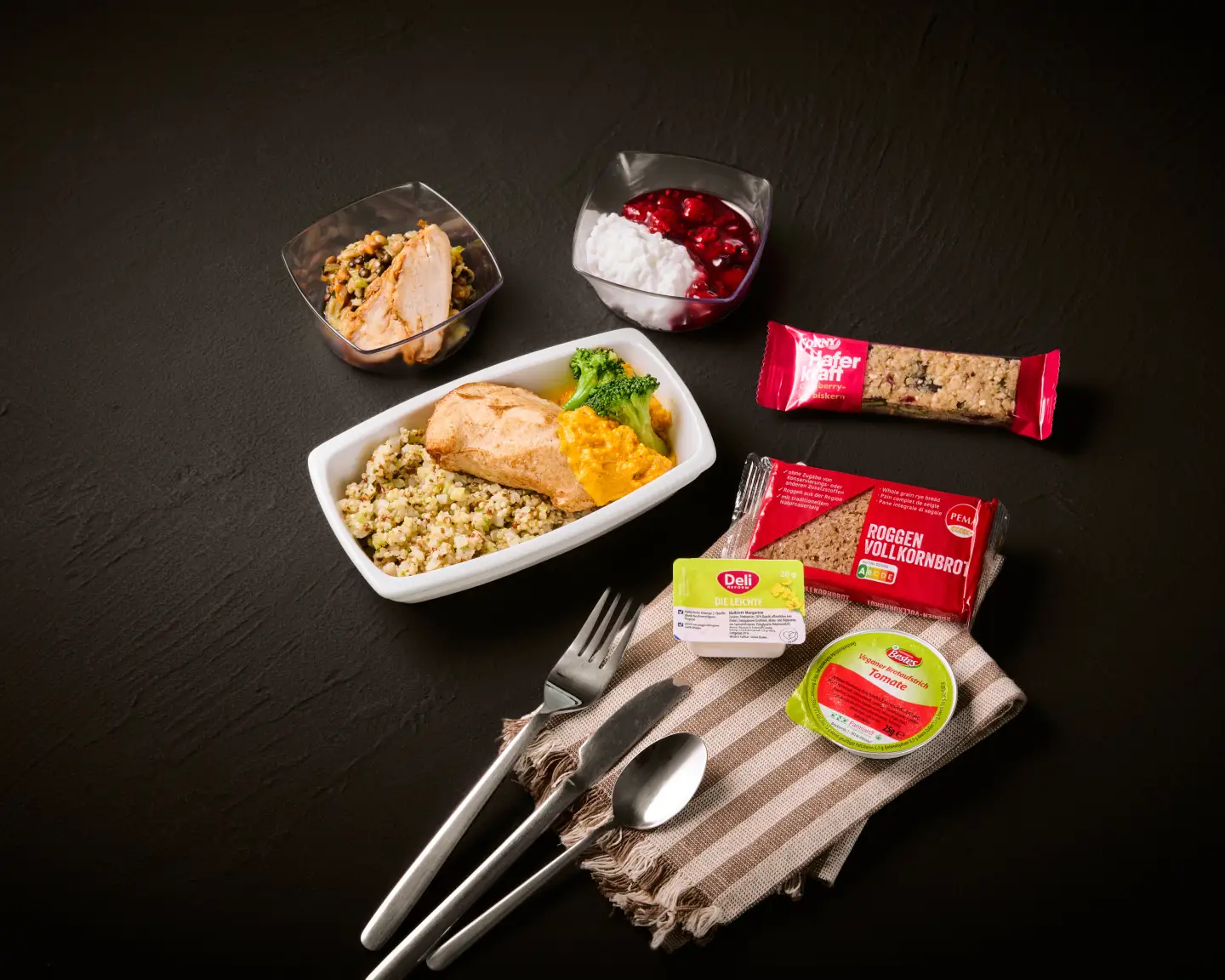 Een maaltijd aan boord met kipfilet, een bijgerecht van kikkererwten, een bijsalade, een mueslireep en een portie smeersel, gepresenteerd tegen een donkere achtergrond.