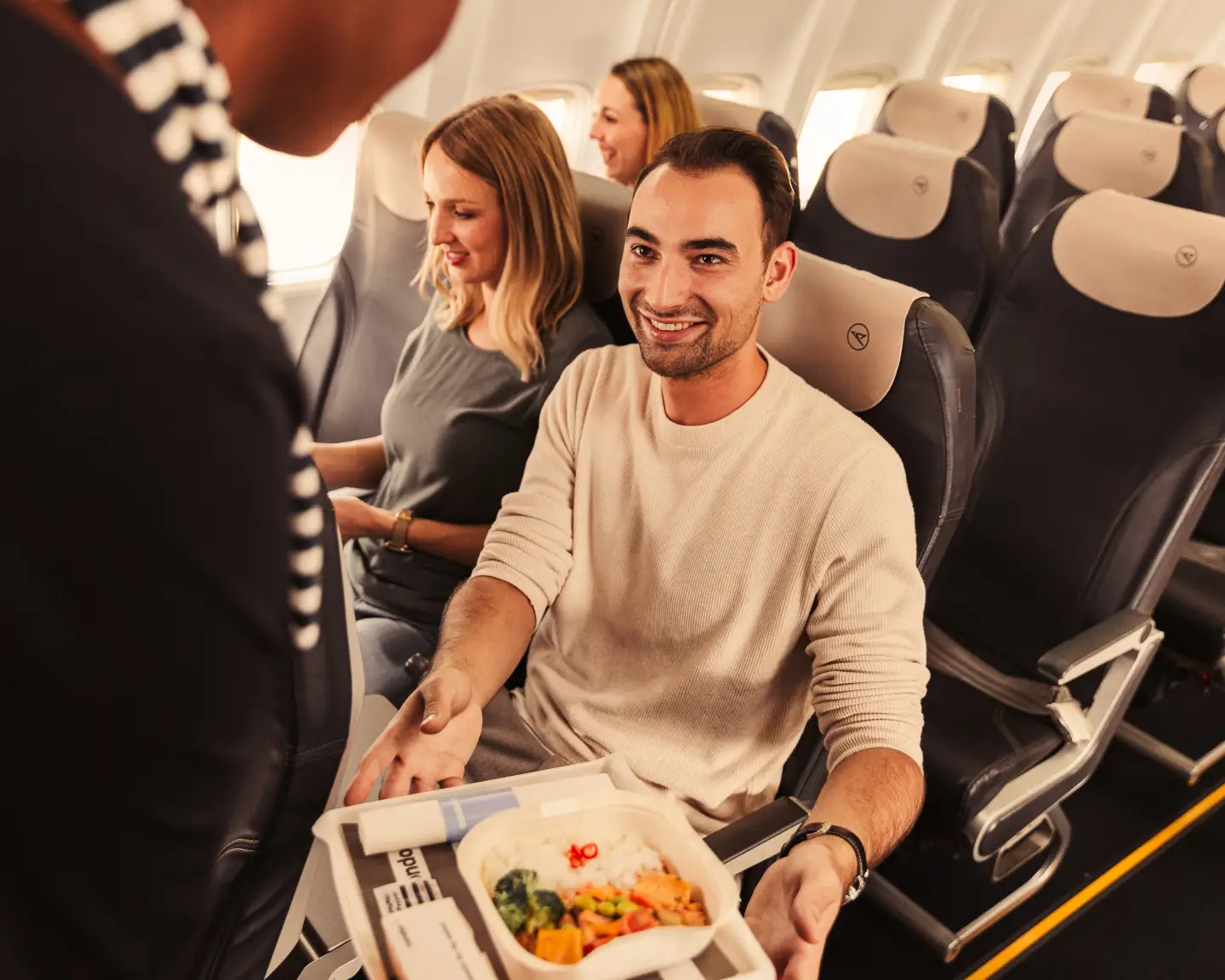 Un hombre sonriente está recibiendo su comida de una auxiliar de vuelo a bordo de un avión de Condor. Al fondo se ven más pasajeros.
