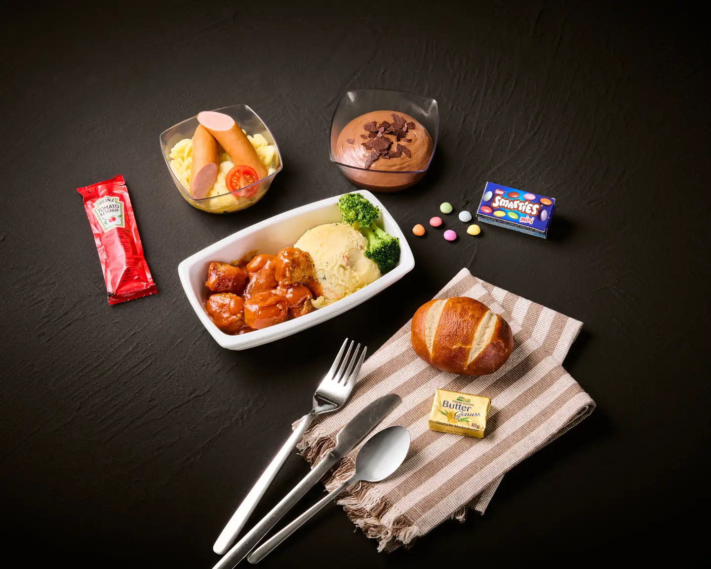 Ilustración de un menú infantil sobre un fondo oscuro, que incluye carne con puré de patatas y brócoli, salchichas con ensalada de patatas, mousse de chocolate, un panecillo y unos Smarties 