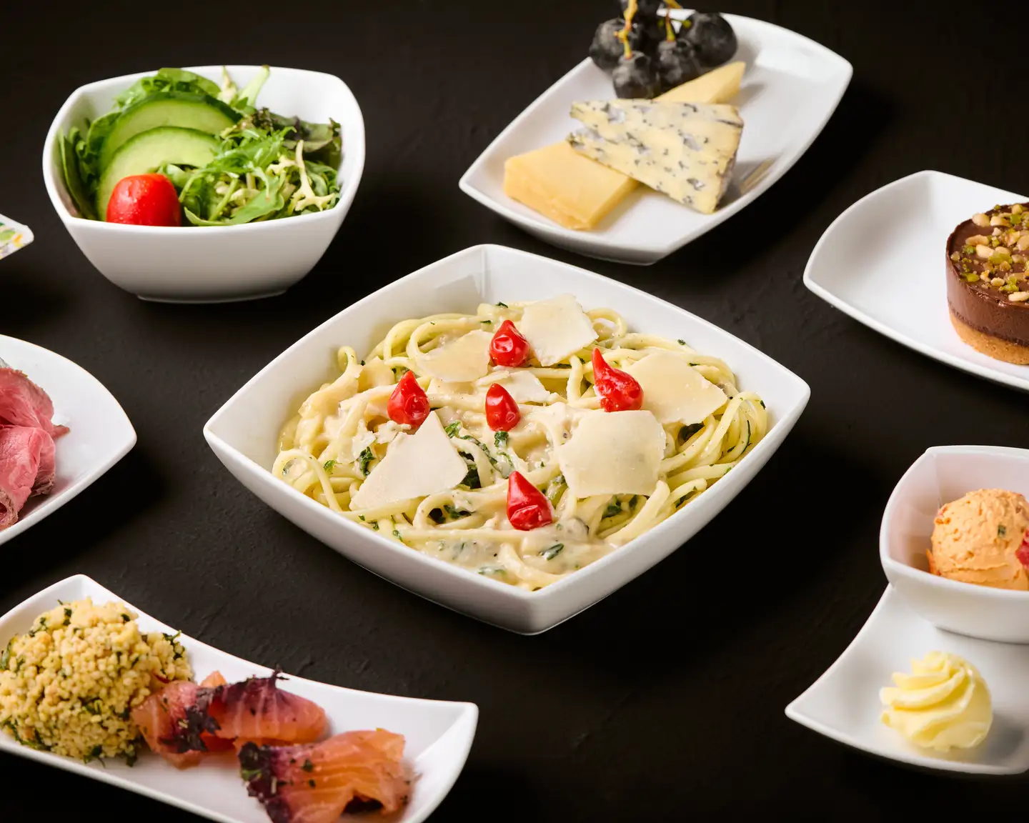 Una comida elegante que consiste en pasta con salsa cremosa, adornada con queso parmesano y tomates cherry rojos, acompañada de una selección de quesos y una ensalada con aguacate sobre un fondo oscuro.