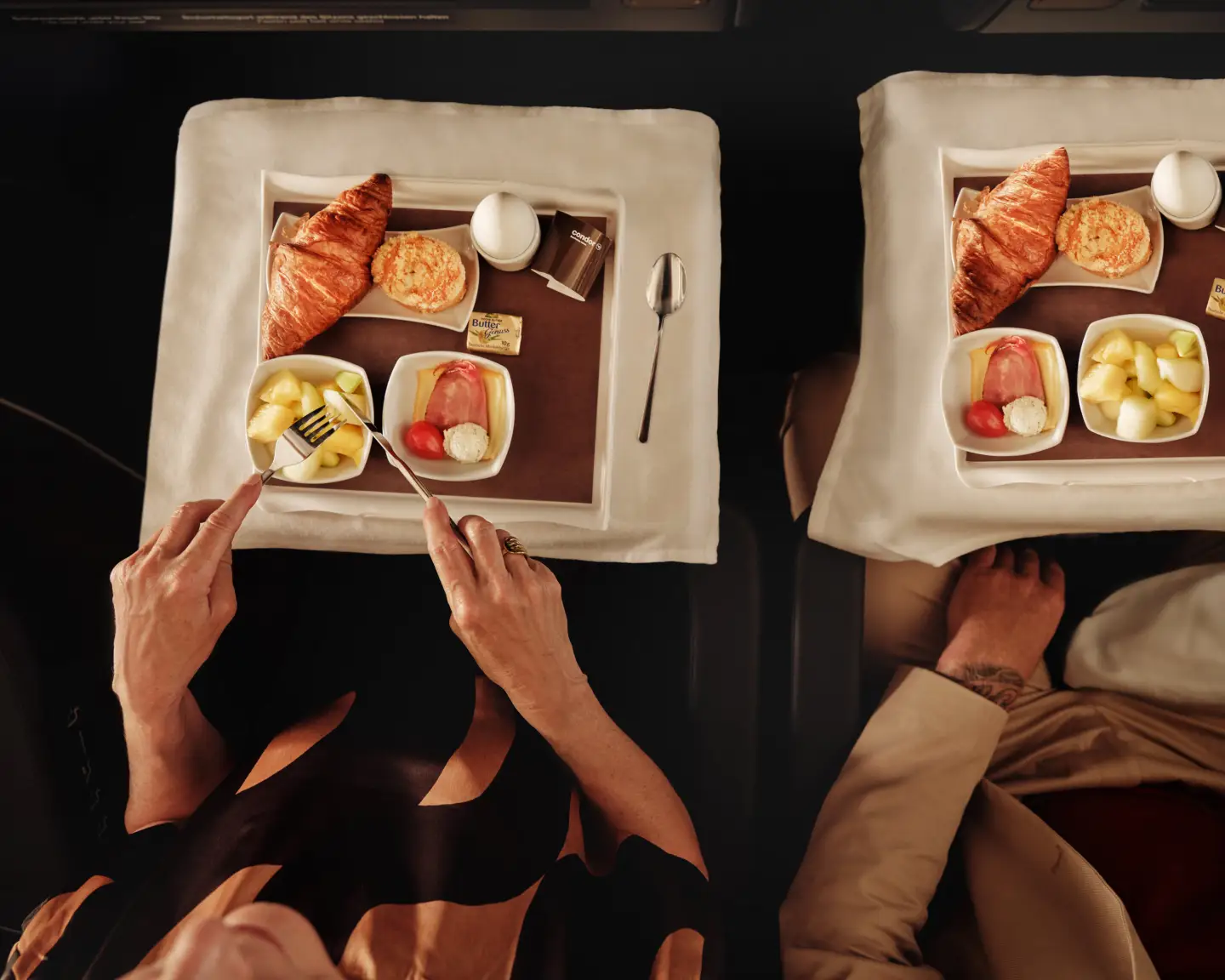 Een koppel aan boord van een vliegtuig geniet van een ontbijt met croissants, eieren, fruit en kaas op elegant gerangschikte dienbladen, met een wit tafelkleed en porseleinen servies.