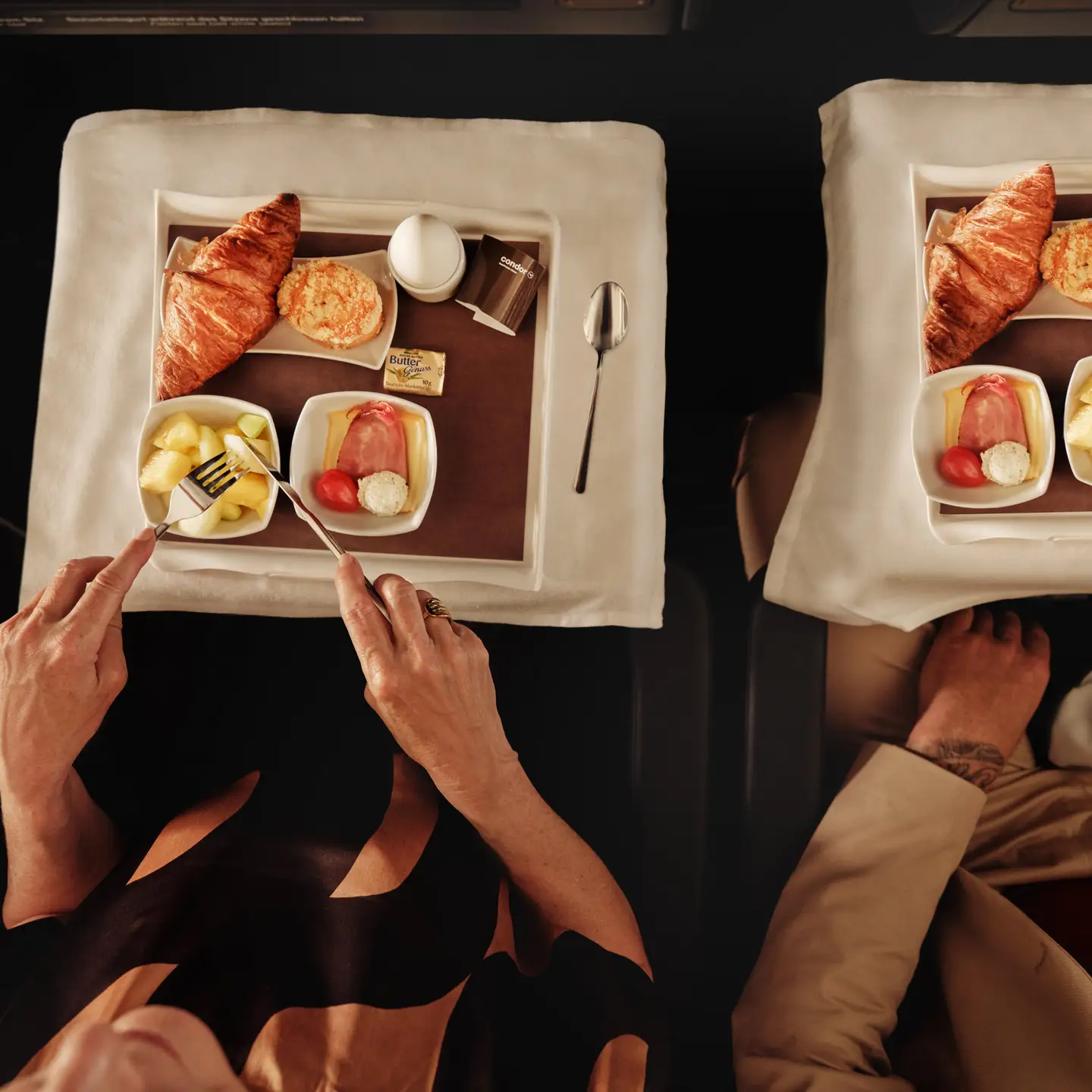 Una pareja a bordo de un avión disfruta de un desayuno que consiste en croissants, huevos, fruta y queso en bandejas elegantemente dispuestas con mantel blanco y vajilla de porcelana.