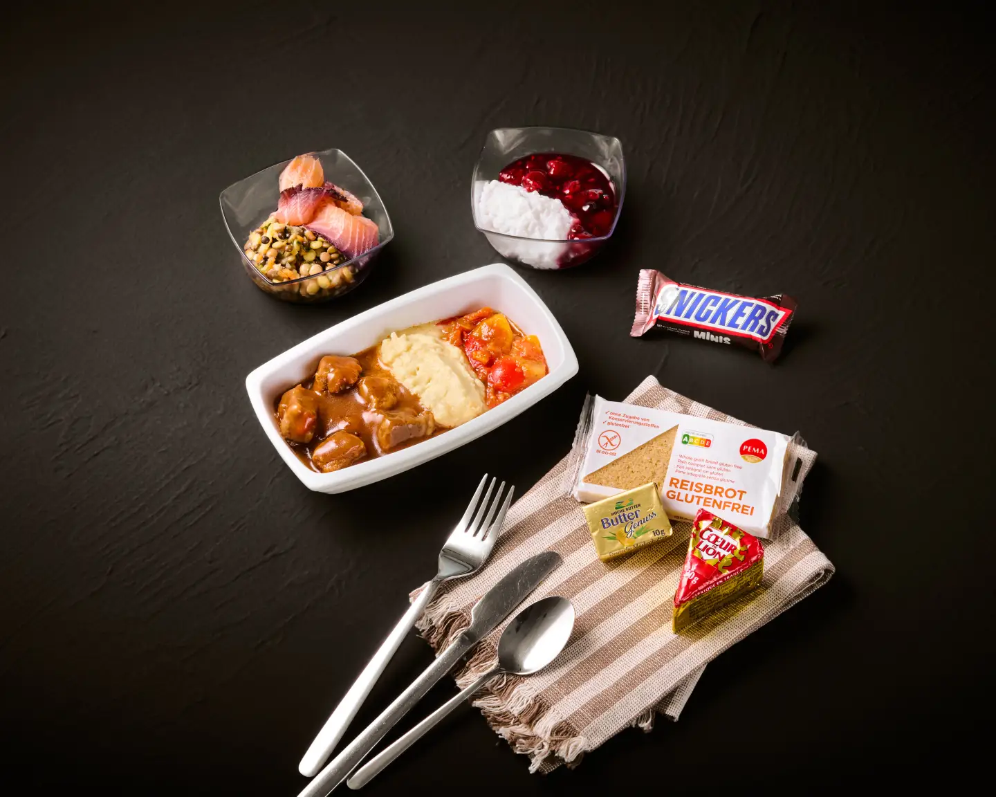 Een maaltijd met verschillende gerechten zoals vlees, aardappelpuree en groenten, plus bijgerechten zoals yoghurt, een mueslireep en een chocoladereep tegen een donkere achtergrond
