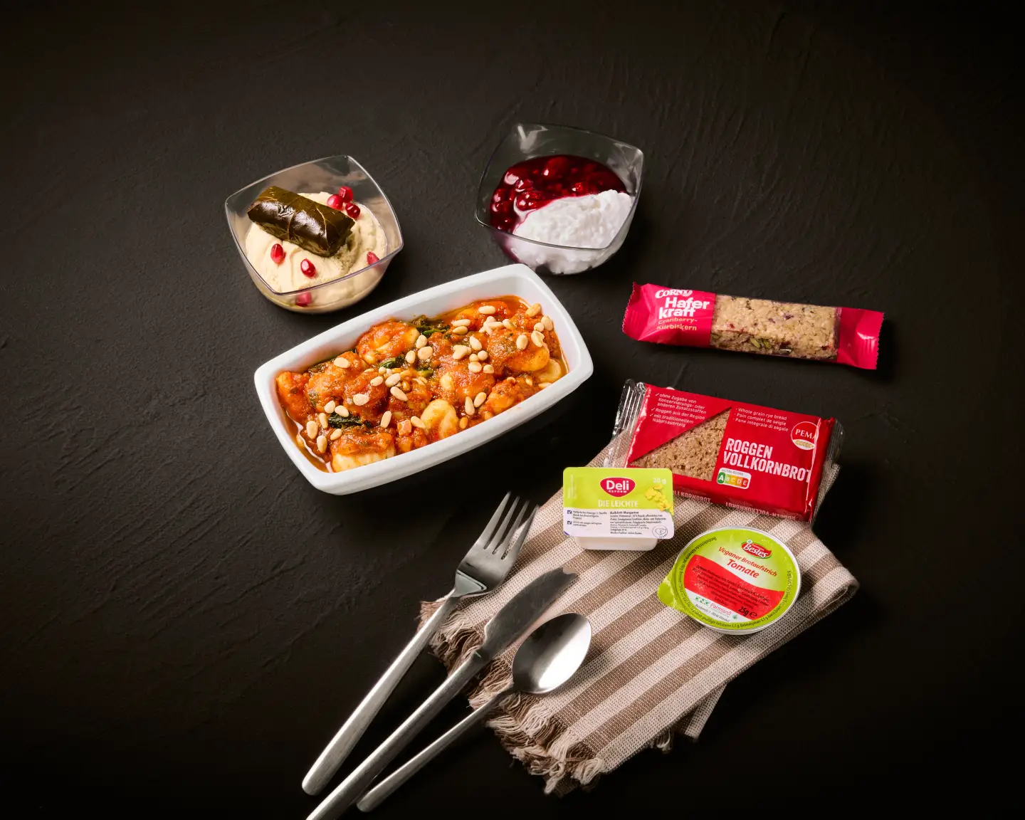 Una comida servida en un avión que incluye un plato de ñoquis con verduras, una guarnición, un postre, una barrita de muesli y un pancito.