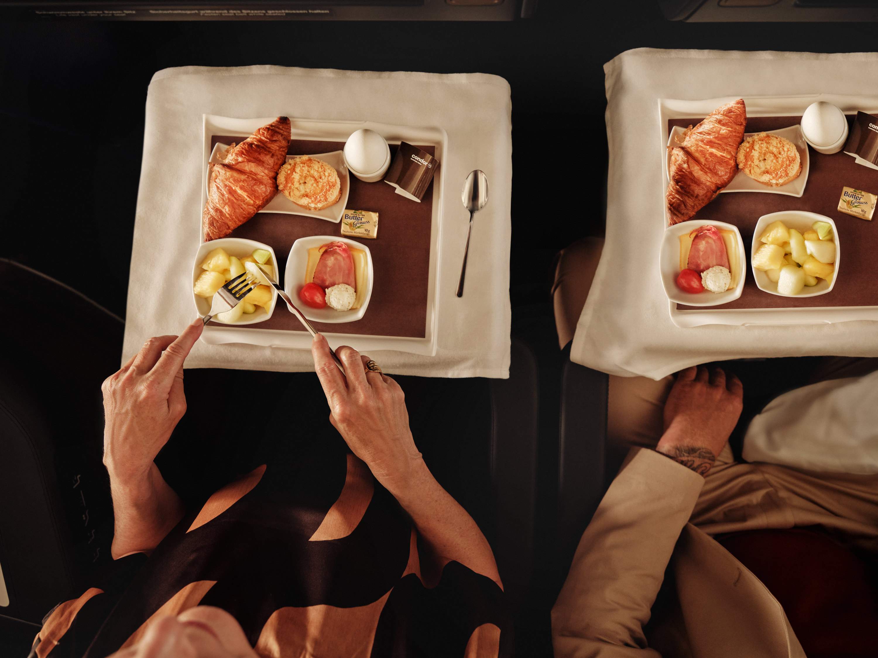 Una pareja a bordo de un avión disfruta de un desayuno que consiste en croissants, huevos, fruta y queso en bandejas elegantemente dispuestas con mantel blanco y vajilla de porcelana.