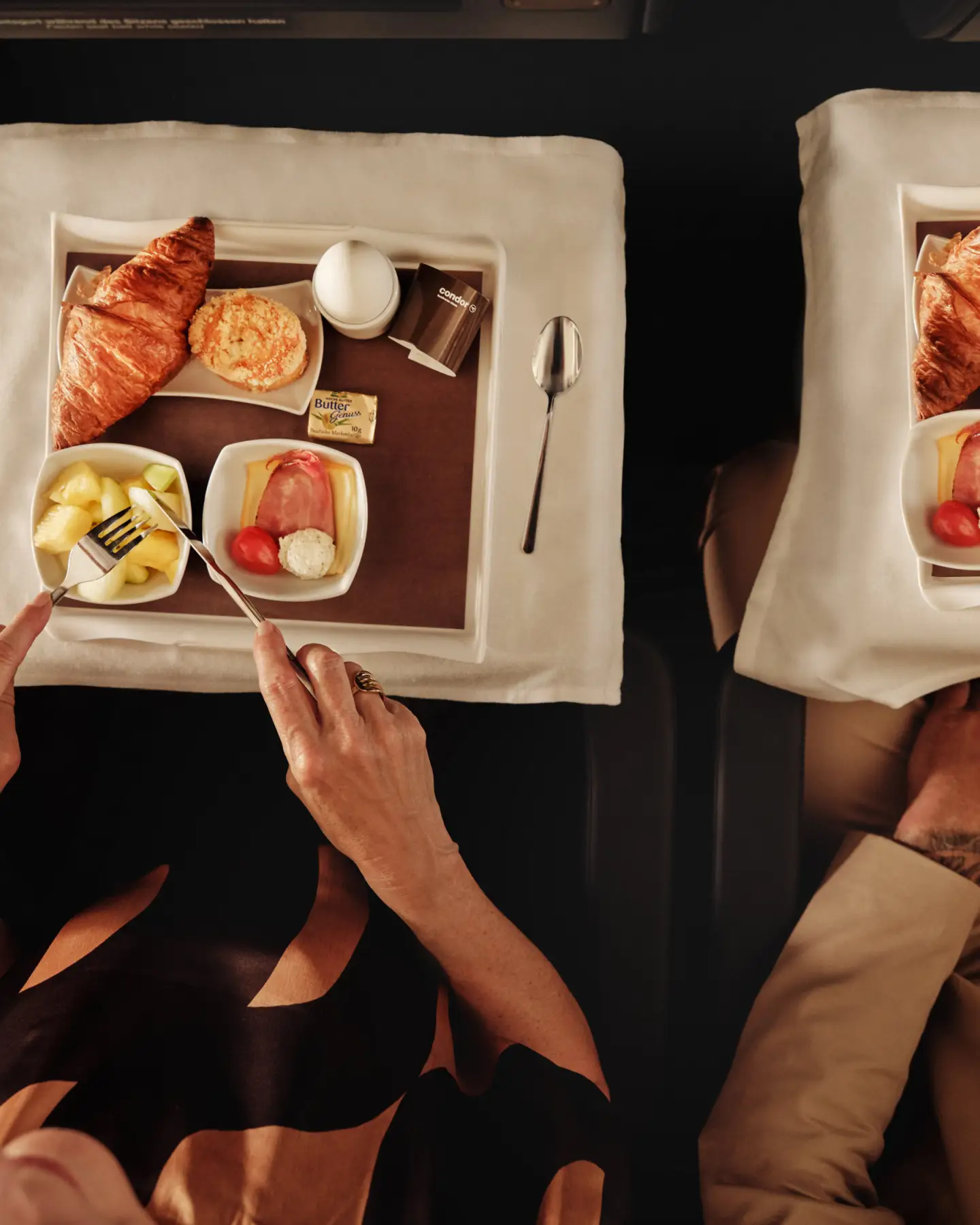 Egy pár a repülőgép fedélzetén élvezi reggelijét, amely croissant-ból, tojásból, gyümölcsből és sajtból áll, elegánsan elrendezett tálcákon, fehér terítővel és porcelán étkészlettel.