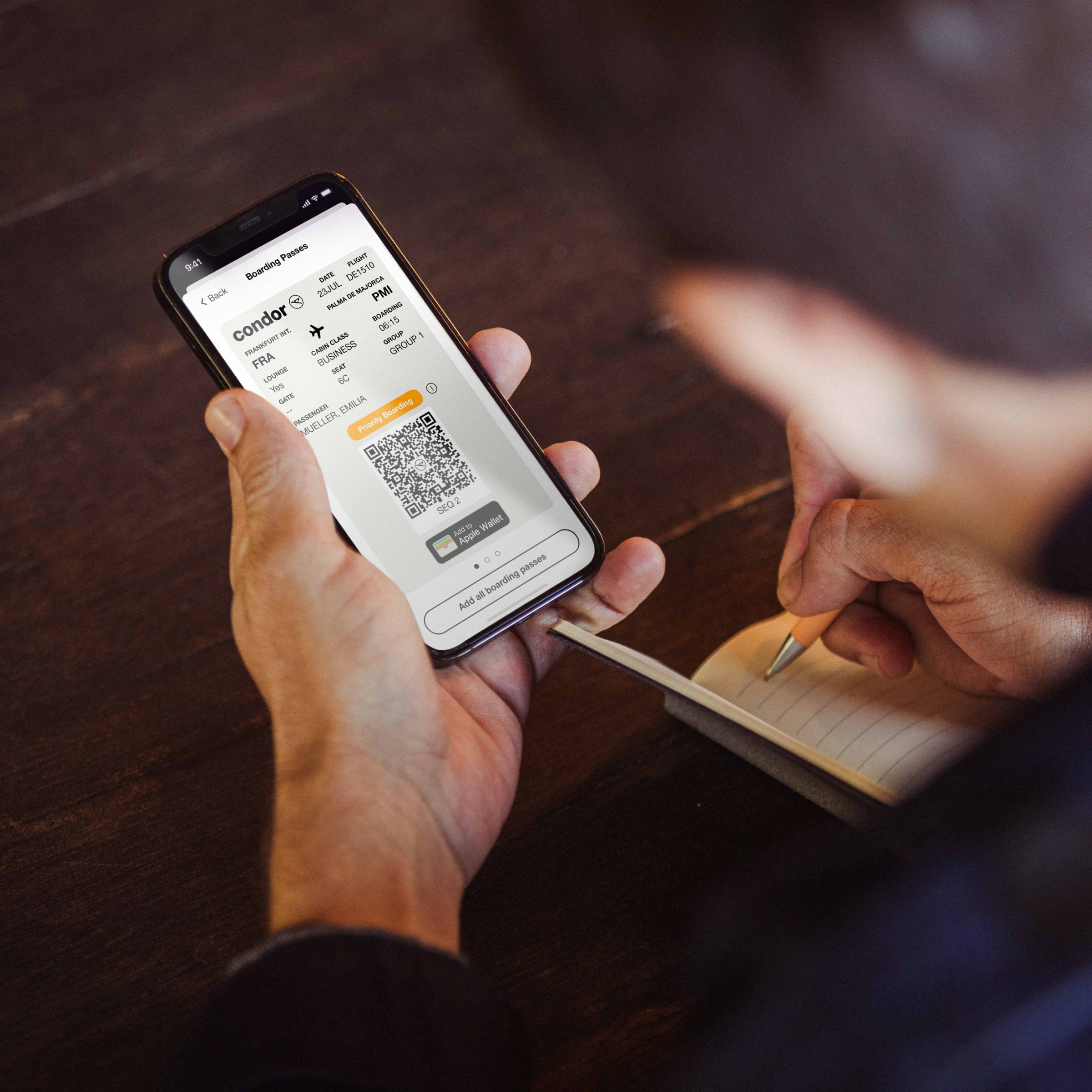 Una persona sostiene un smartphone en el que se muestra una tarjeta de embarque digital para un vuelo de Condor, mientras escribe notas en un cuaderno con un bolígrafo.