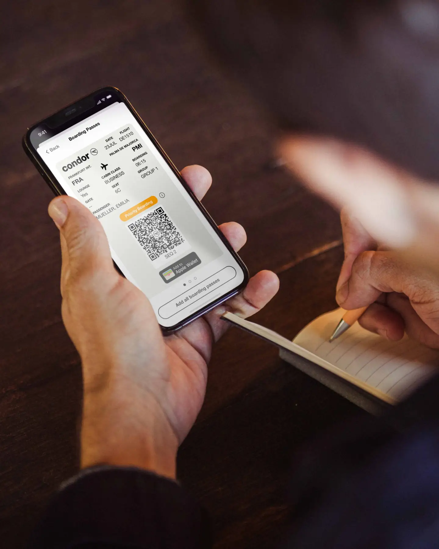 Una persona sostiene un smartphone en el que se muestra una tarjeta de embarque digital para un vuelo de Condor, mientras escribe notas en un cuaderno con un bolígrafo.
