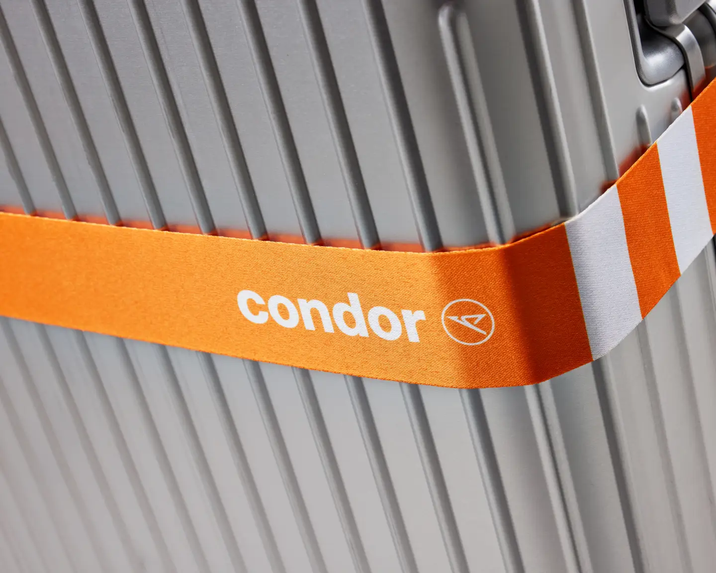 Grande plano de uma correia de bagagem Condor cor de laranja e branca presa a uma mala prateada.